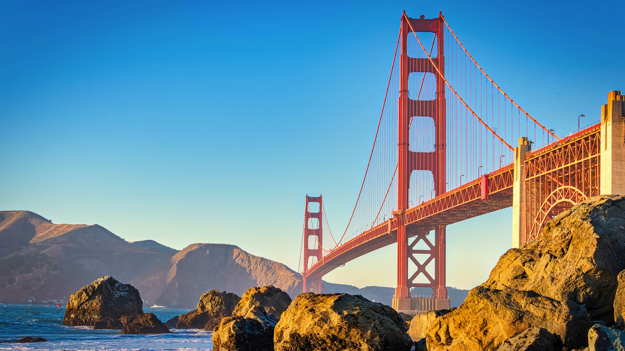 Rote Hängebrücke bei sonnigem Wetter über einem blauen Meer mit Felsen im Vordergrund.