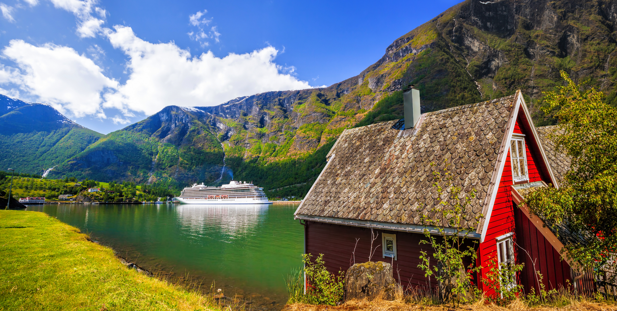 Rotes Haus am Fjord mit Kreuzfahrtschiff und Bergen im Hintergrund bei Sonnenschein.