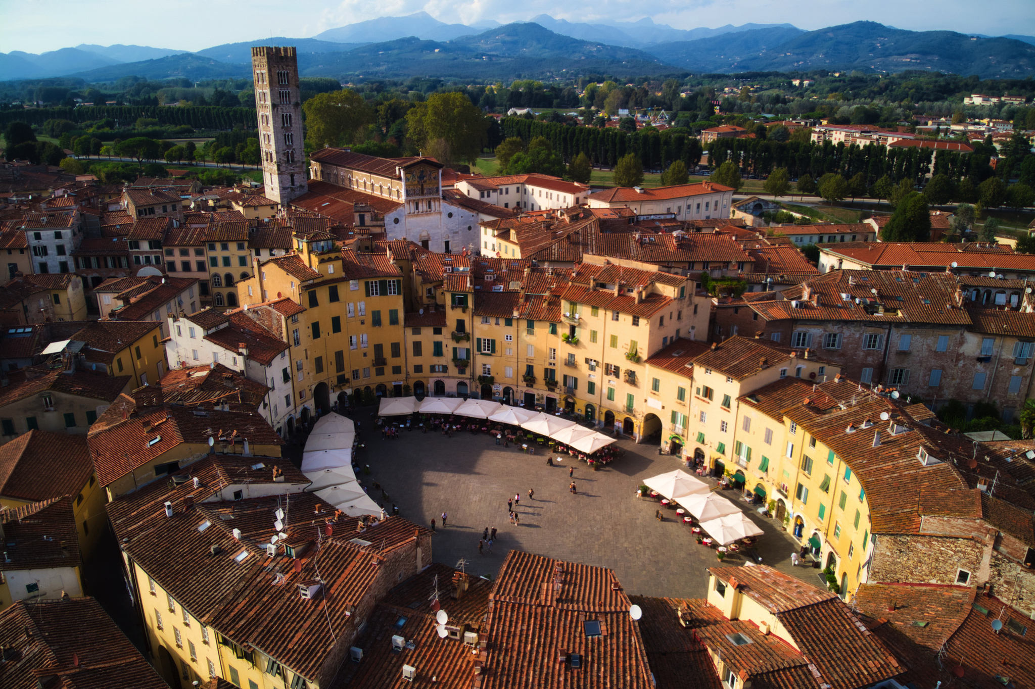 Amphitheaterplatz in Lucca Luftaufnahme eines kreisförmigen Platzes mit umliegenden Gebäuden und Bergen im Hintergrund.
