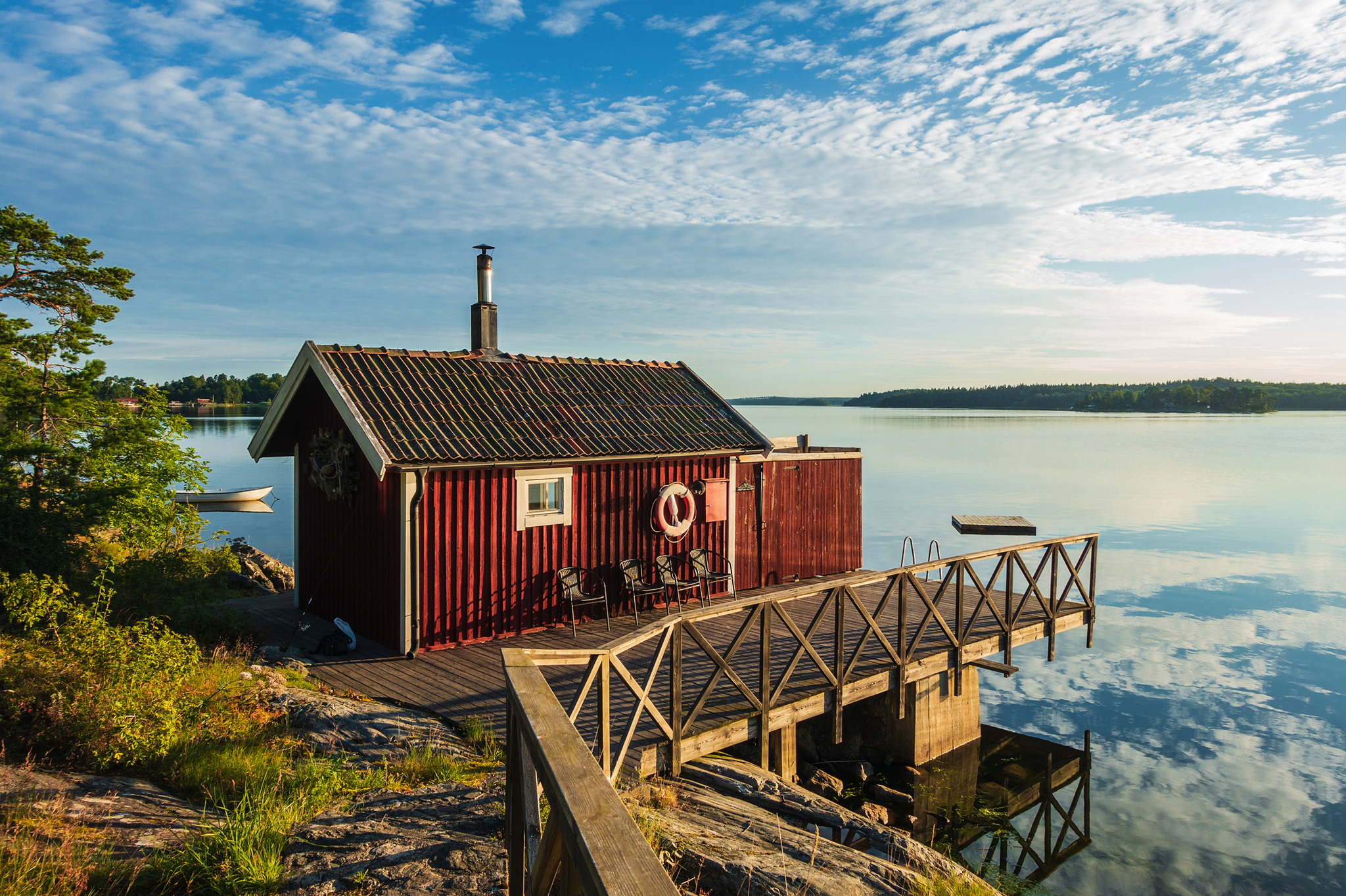 Kleines rotes Ferienhaus mit Holzsteg am Wasser im Stockholmer Schärengarten bei ruhiger See und blauem Himmel.