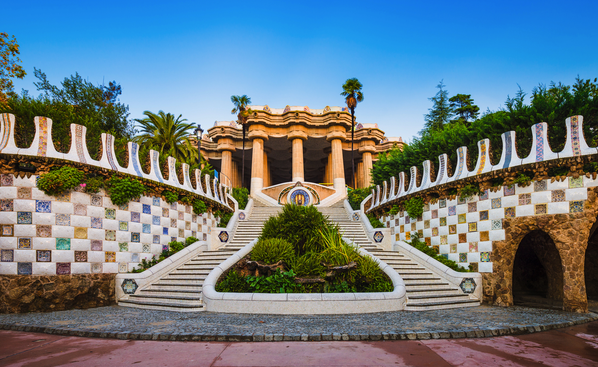 Der Park Güell in Barcelona mit einzigartiger, bunter Architektur und geschwungenen Formen entworfen von Antoni Gaudí, umgeben von Pflanzen und Palmen.
