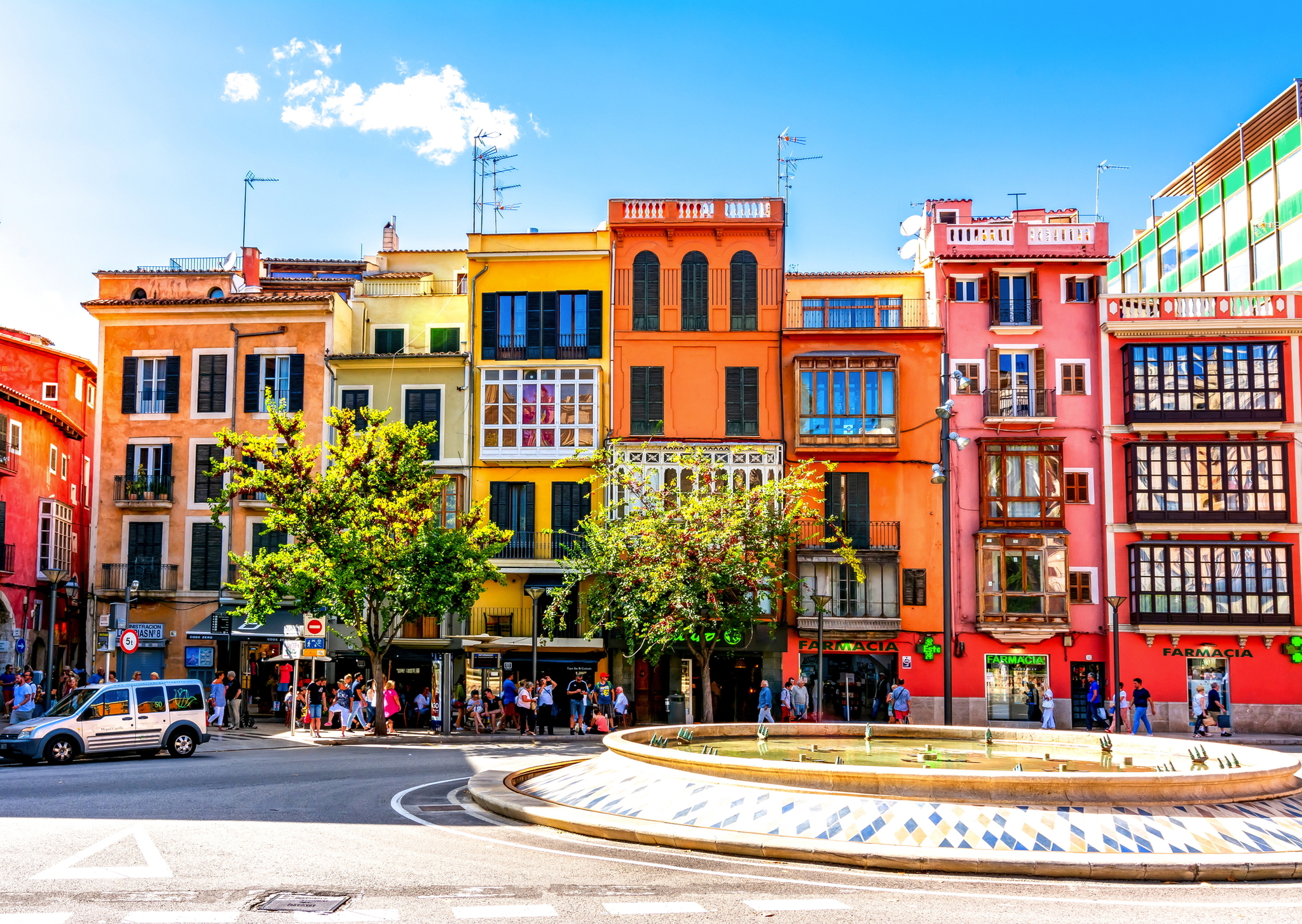 Bunte Häuserfassaden an belebter Straße mit Brunnen und passierenden Autos bei sonnigem Wetter.