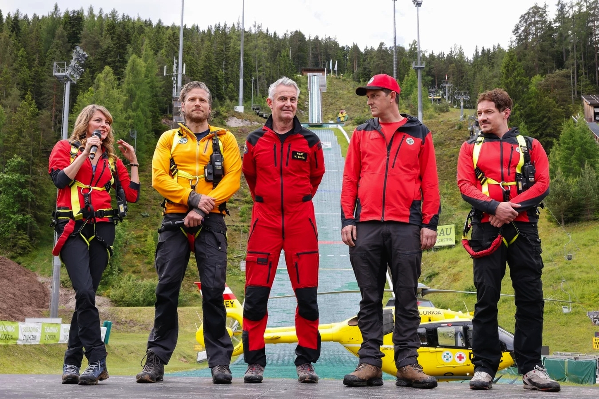 Fünf Personen in Outdoor-Kleidung vor grüner Waldkulisse und Skisprungschanze.