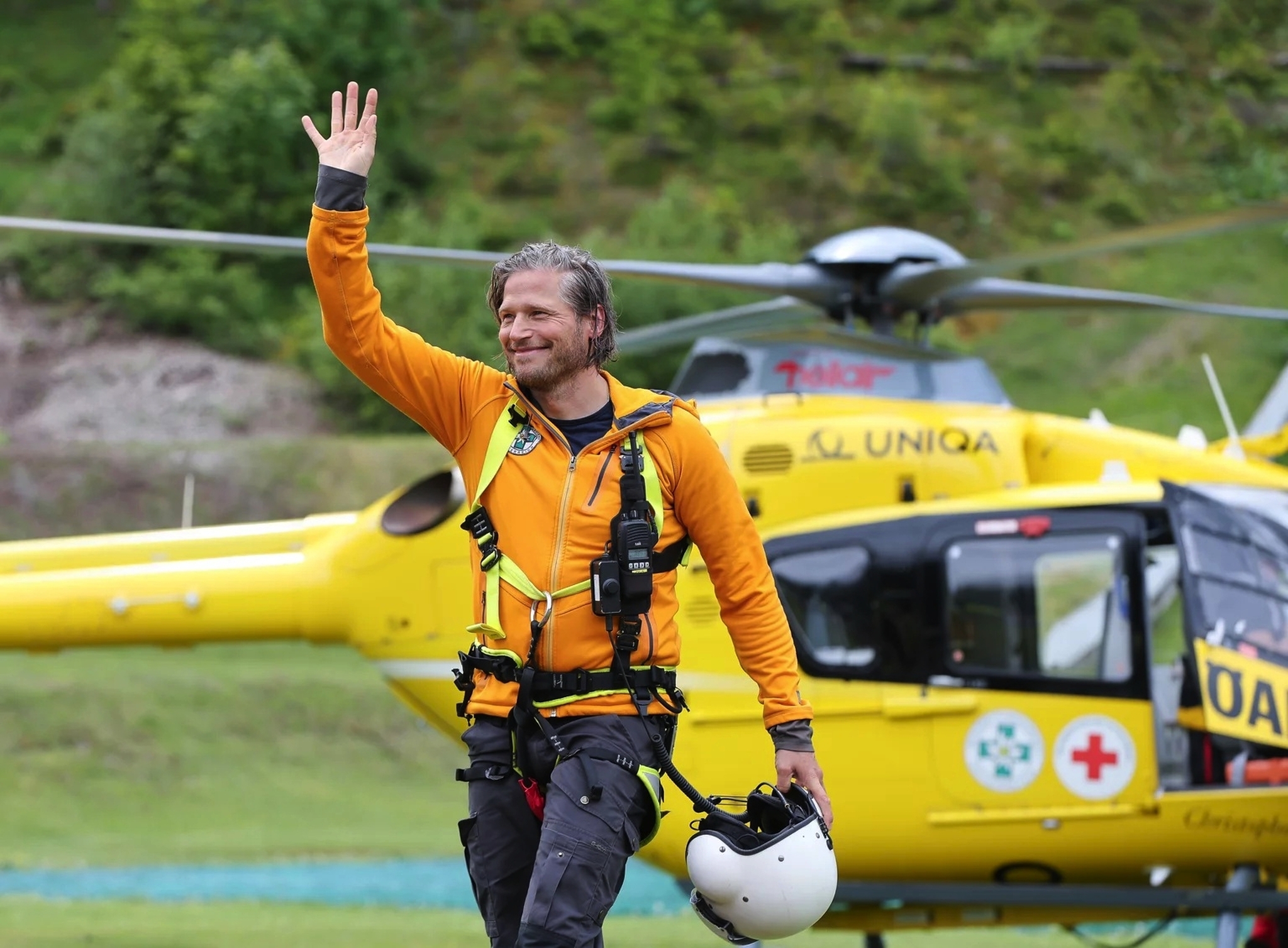 Ein Rettungshelikopter im Hintergrund, ein Mann in orangem Outfit steht im Vordergrund und winkt.
