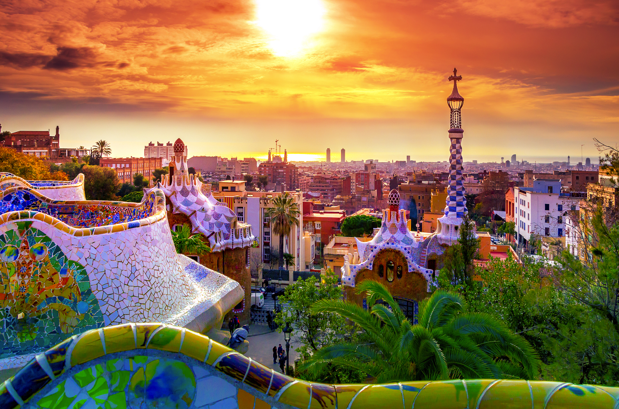 Blick auf die Stadt Barcelona bei Sonnenuntergang vom Park Güell, mit den berühmten bunten Mosaiken und Architektur von Antoni Gaudí im Vordergrund.