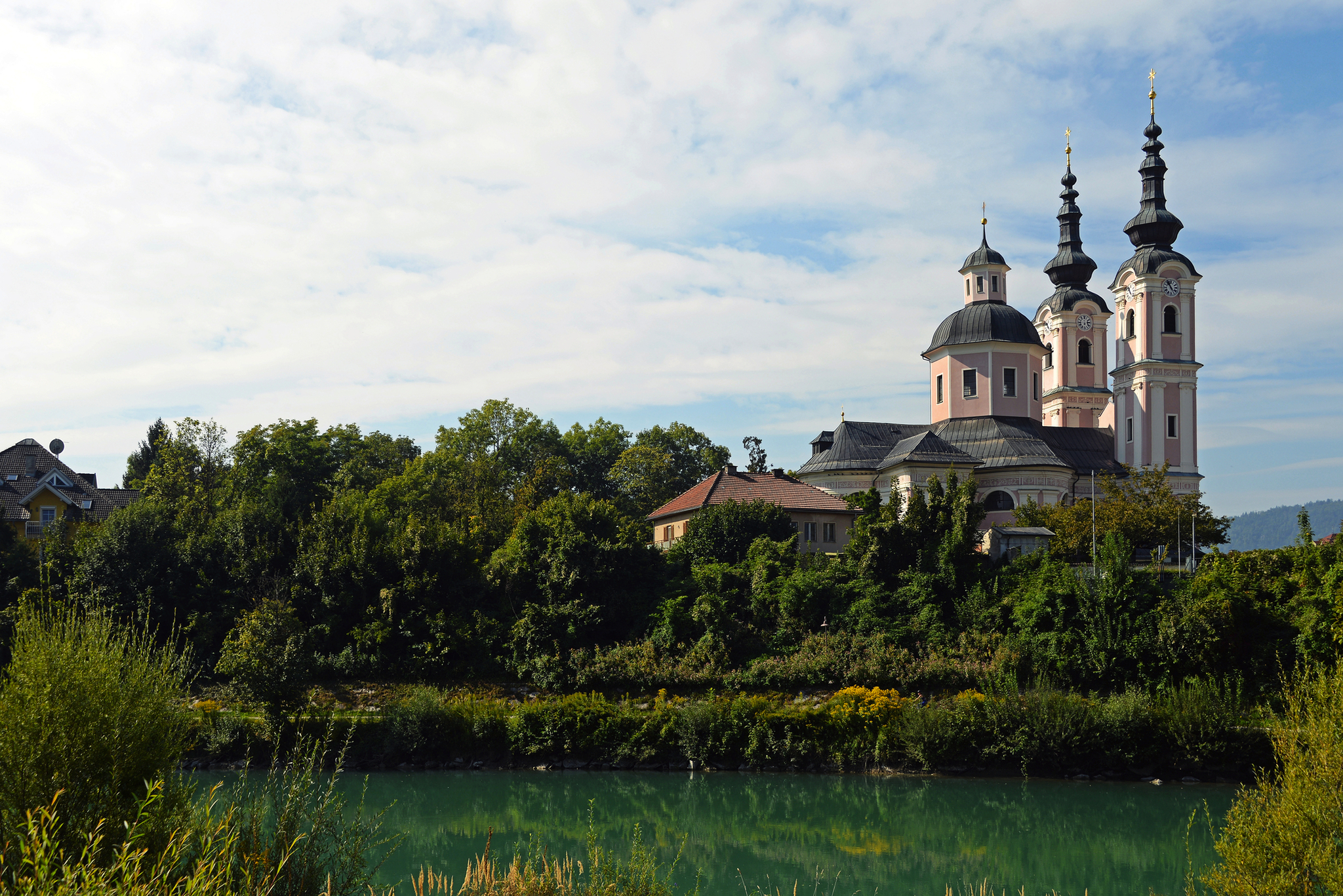 Barockkirche mit Türmen am Flussufer, umgeben von Bäumen und bewölktem Himmel.