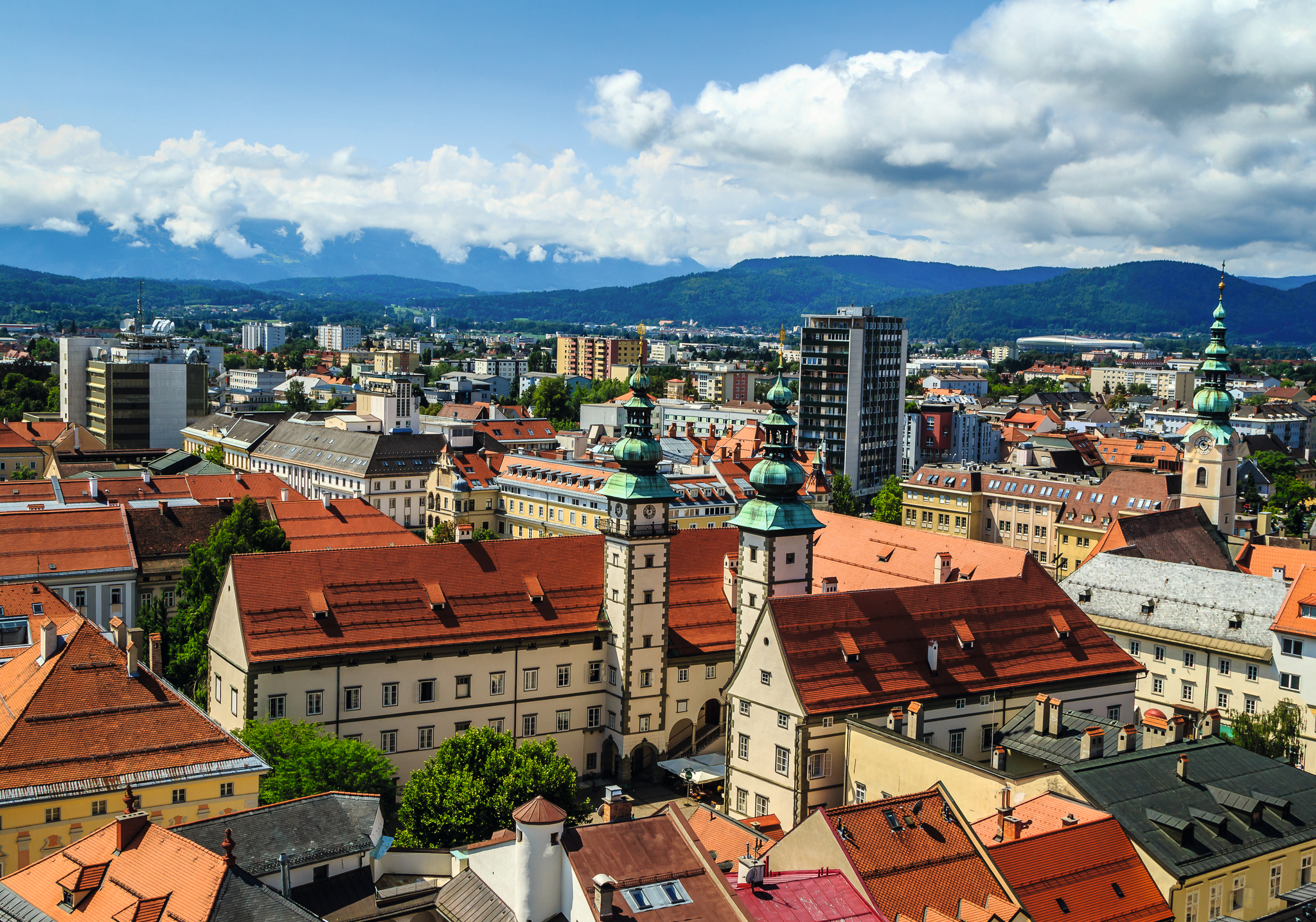 Stadtansicht mit historischen Gebäuden, roten Dächern und Bergen im Hintergrund.