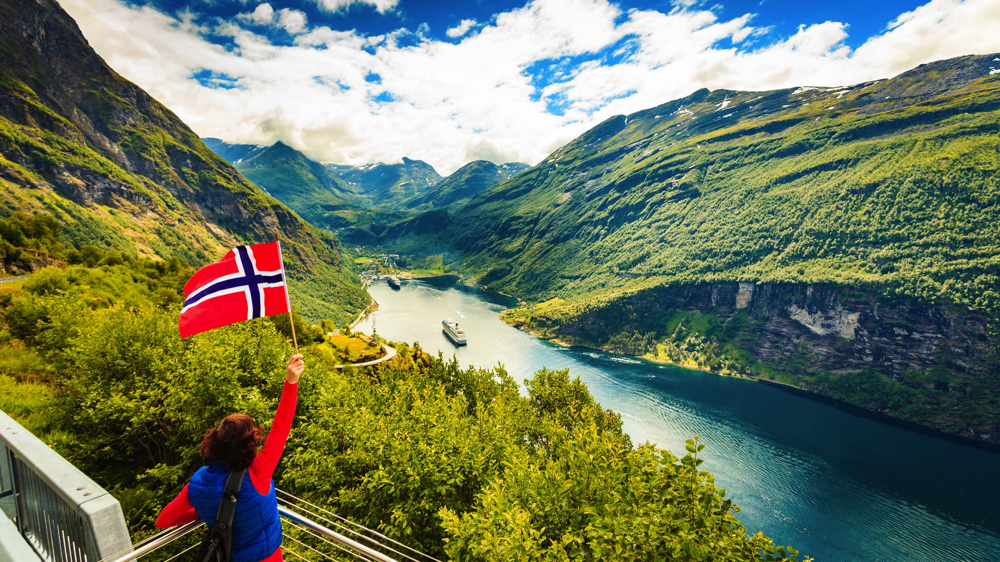 Person schwenkt norwegische Flagge vor Fjordlandschaft.