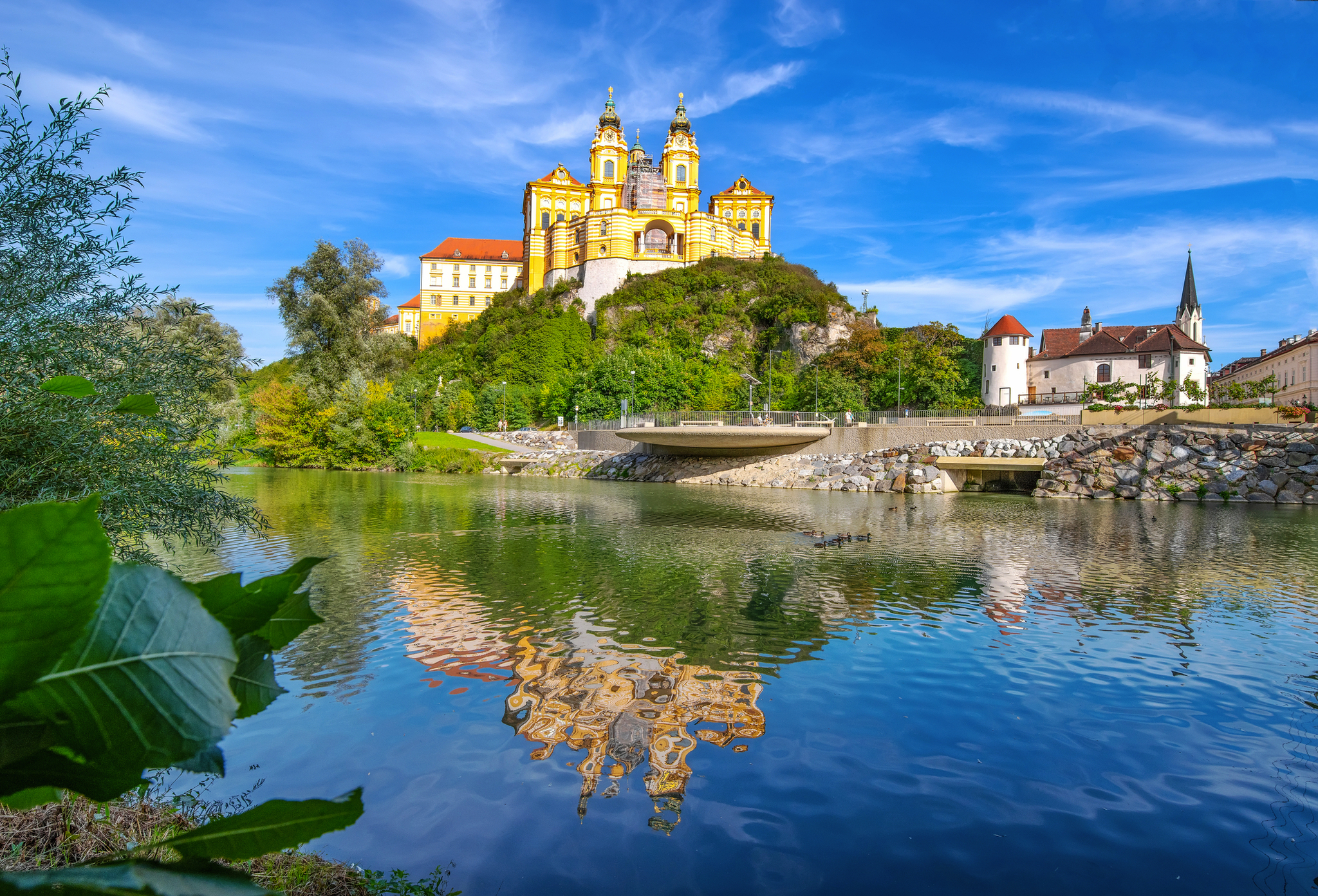 Barockes Schloss auf Hügel spiegelt sich im ruhigen Fluss unter blauem Himmel.