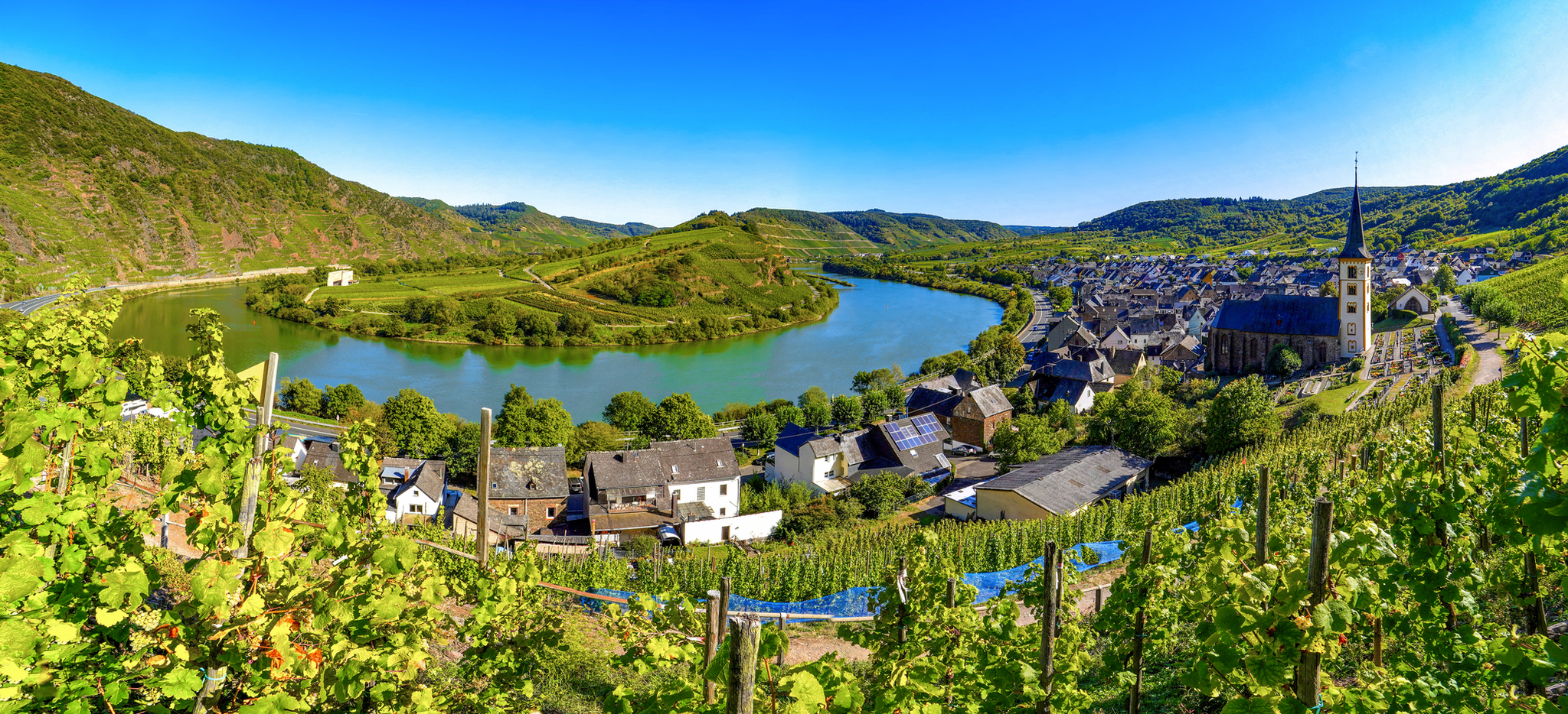 Blick auf malerisches Flusstal mit Dorf und Kirche, umgeben von Hügeln und Weinreben.