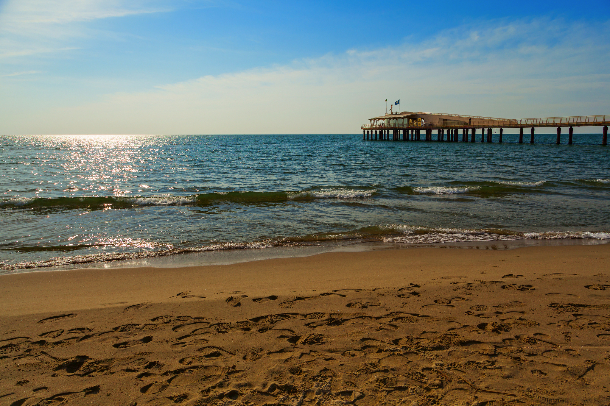 Sonniger Strand mit Fußspuren und Holzsteg am Meer.
