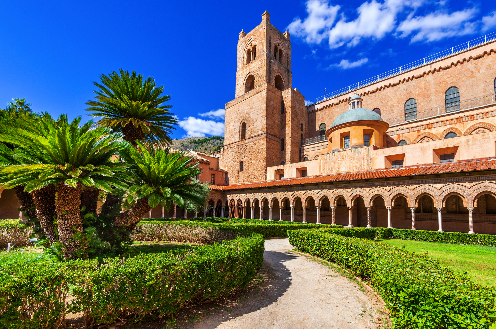 Kreuzgang eines Klosters mit Palmen und historischem Turm unter blauem Himmel.