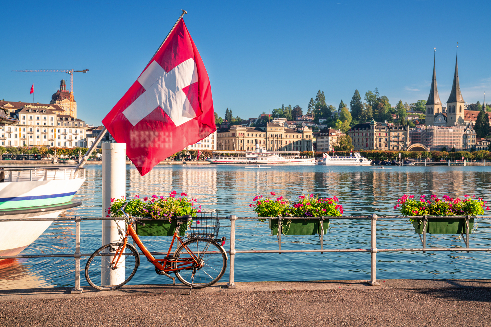 Schweizer Flagge, Fahrrad und blühende Blumen am Seeufer, Stadt im Hintergrund.