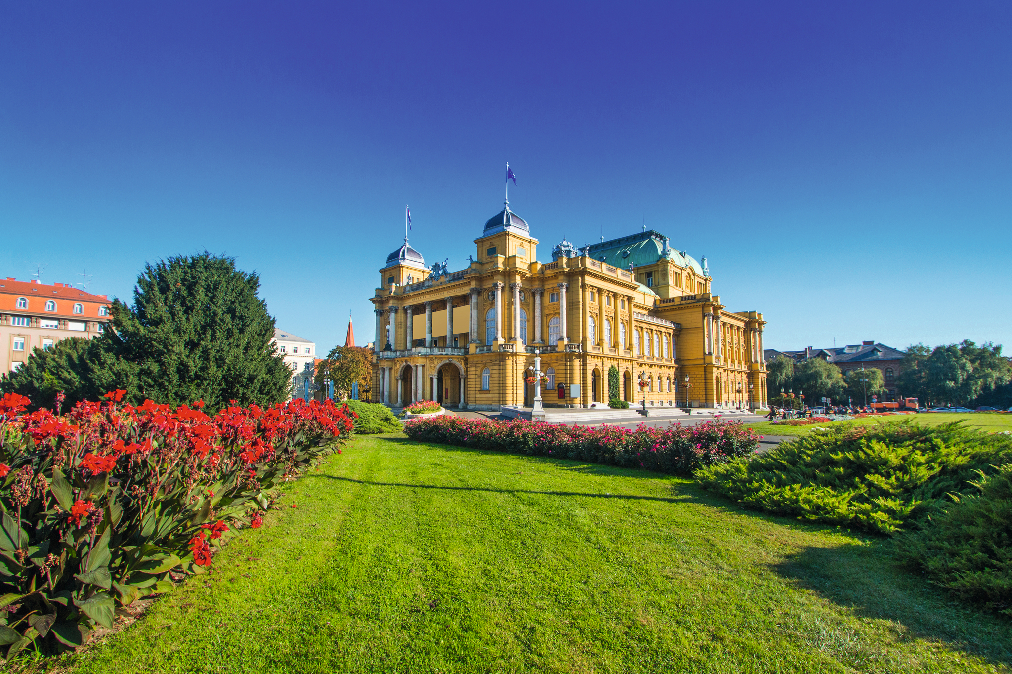 Historisches Gebäude mit Park, blauer Himmel, rote Blumen links im Vordergrund.