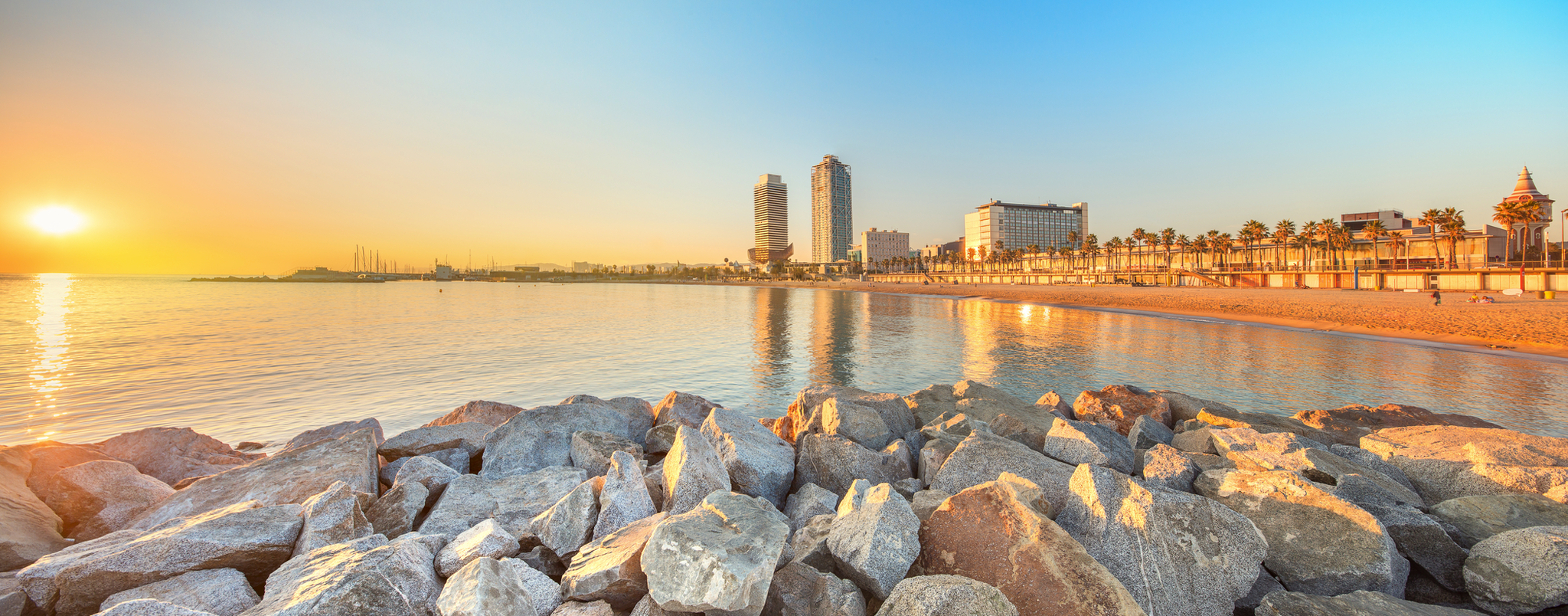 Strand mit Felsen im Vordergrund, Stadt und Sonnenuntergang im Hintergrund.
