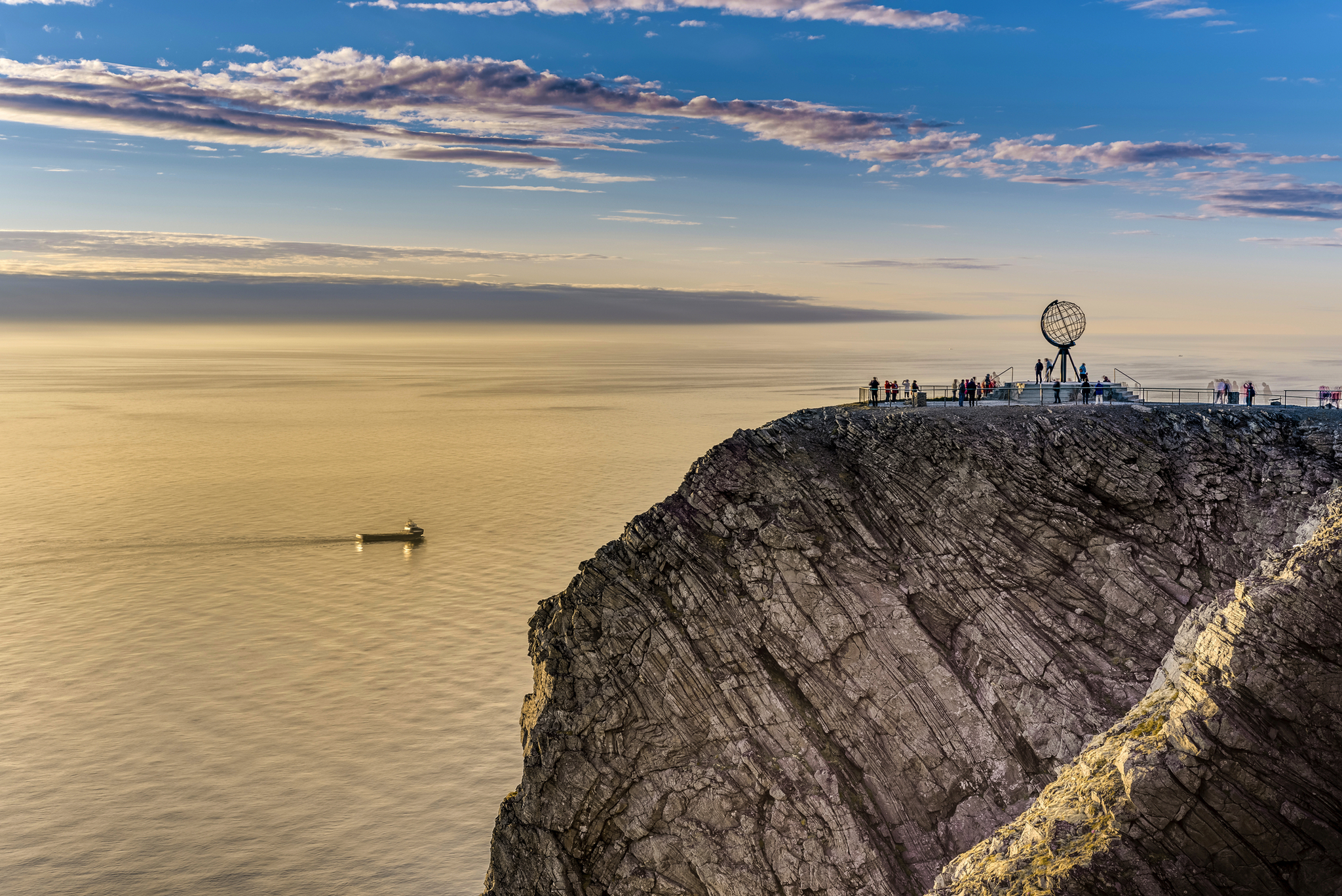 Felsklippe mit Globus-Skulptur am Meer, Leute schauen aufs Wasser.