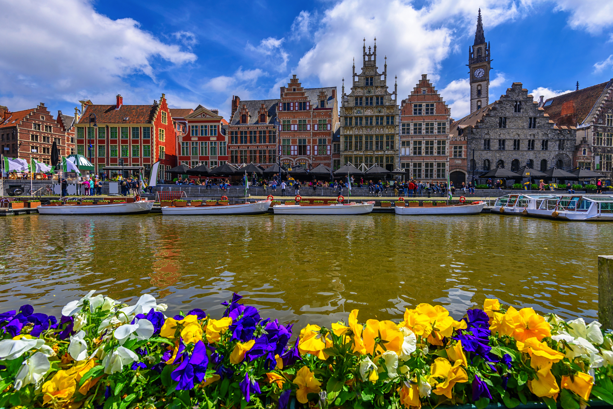 Blumenbeet vor bunten Häusern und Kanal in einer historischen Stadt bei klarem Himmel.