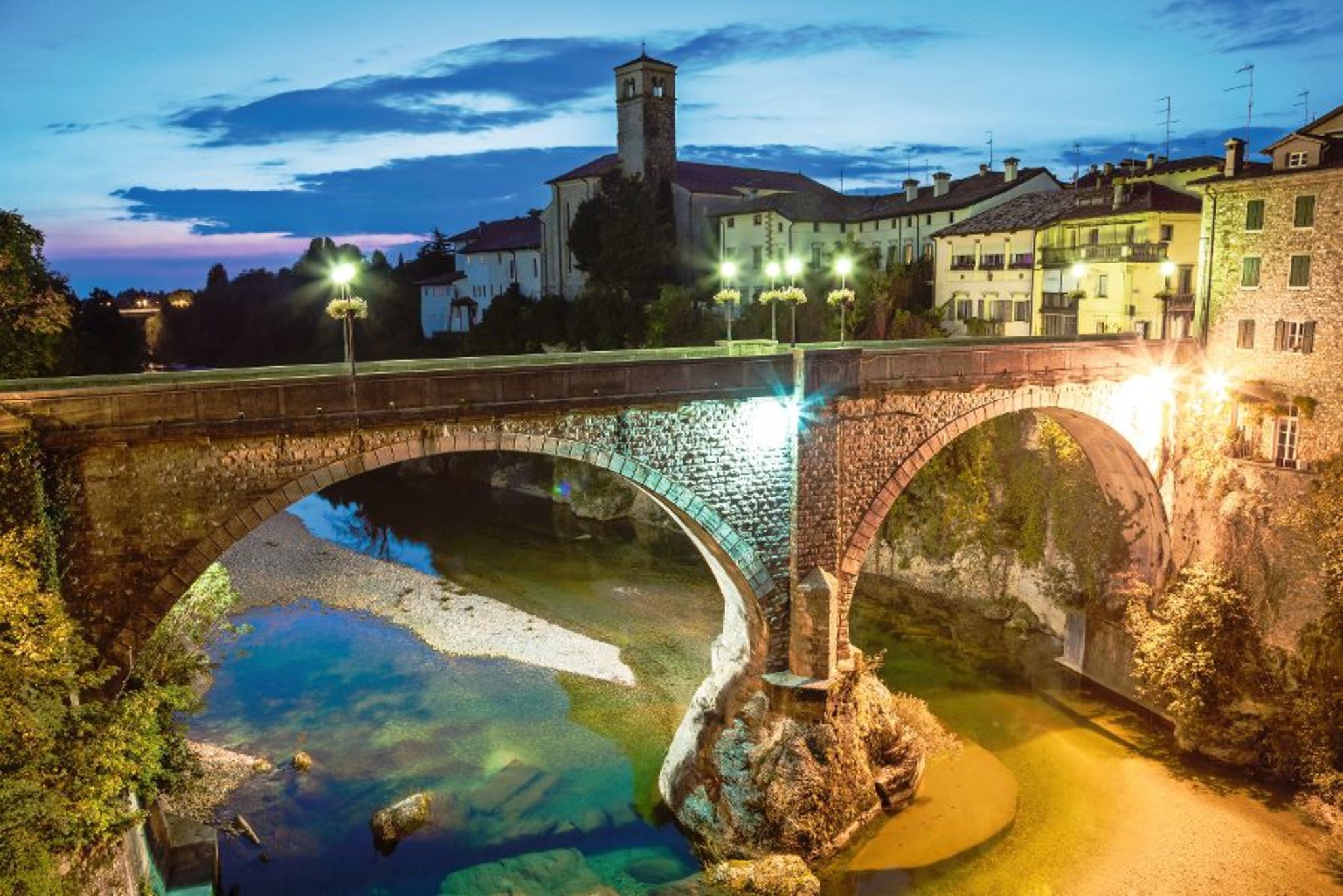 Steinbrücke bei Abenddämmerung über Fluss mit beleuchteten Gebäuden im Hintergrund.