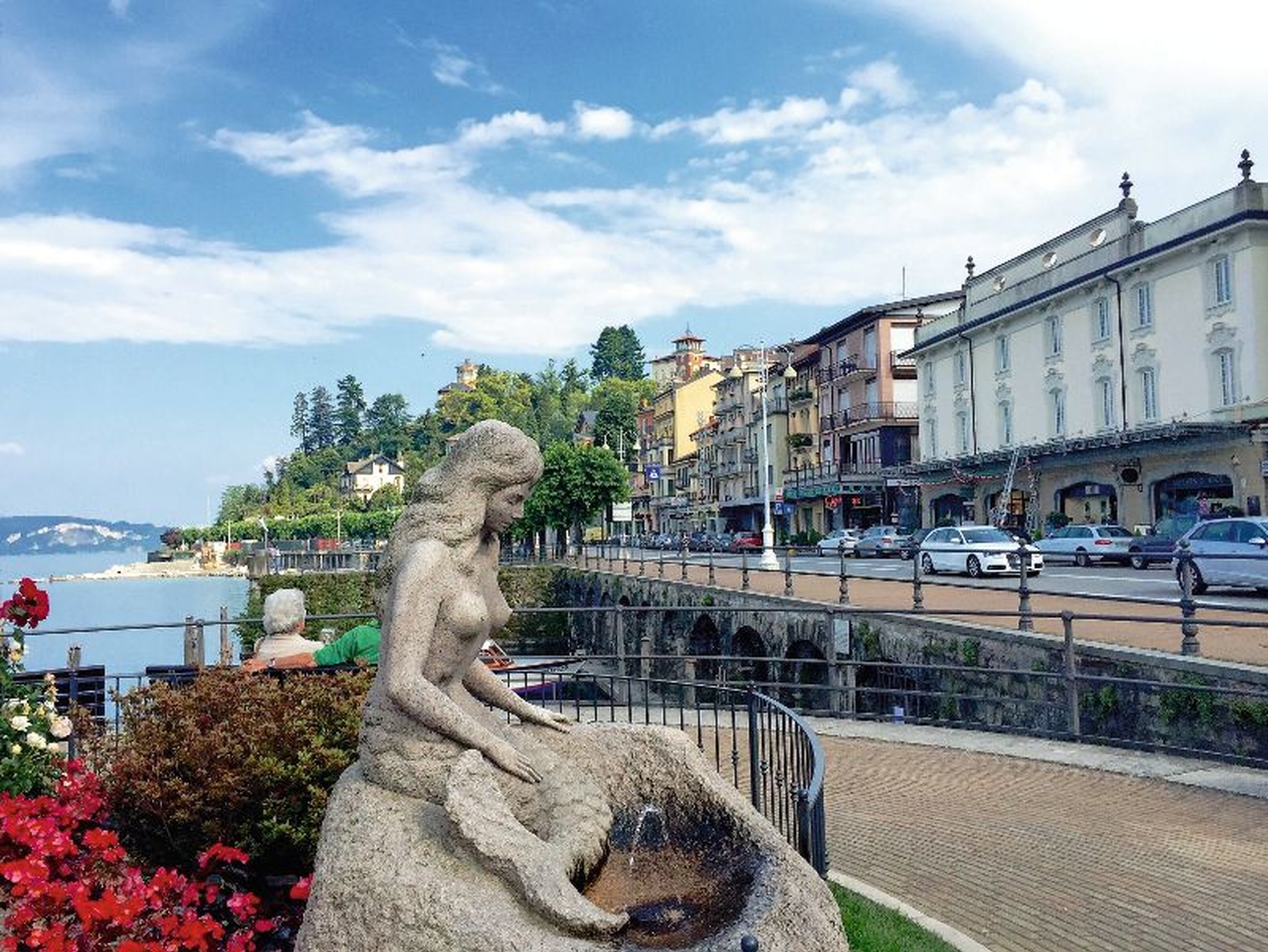 Statue einer Frau am Ufer mit umliegender Architektur und blühenden Blumen.