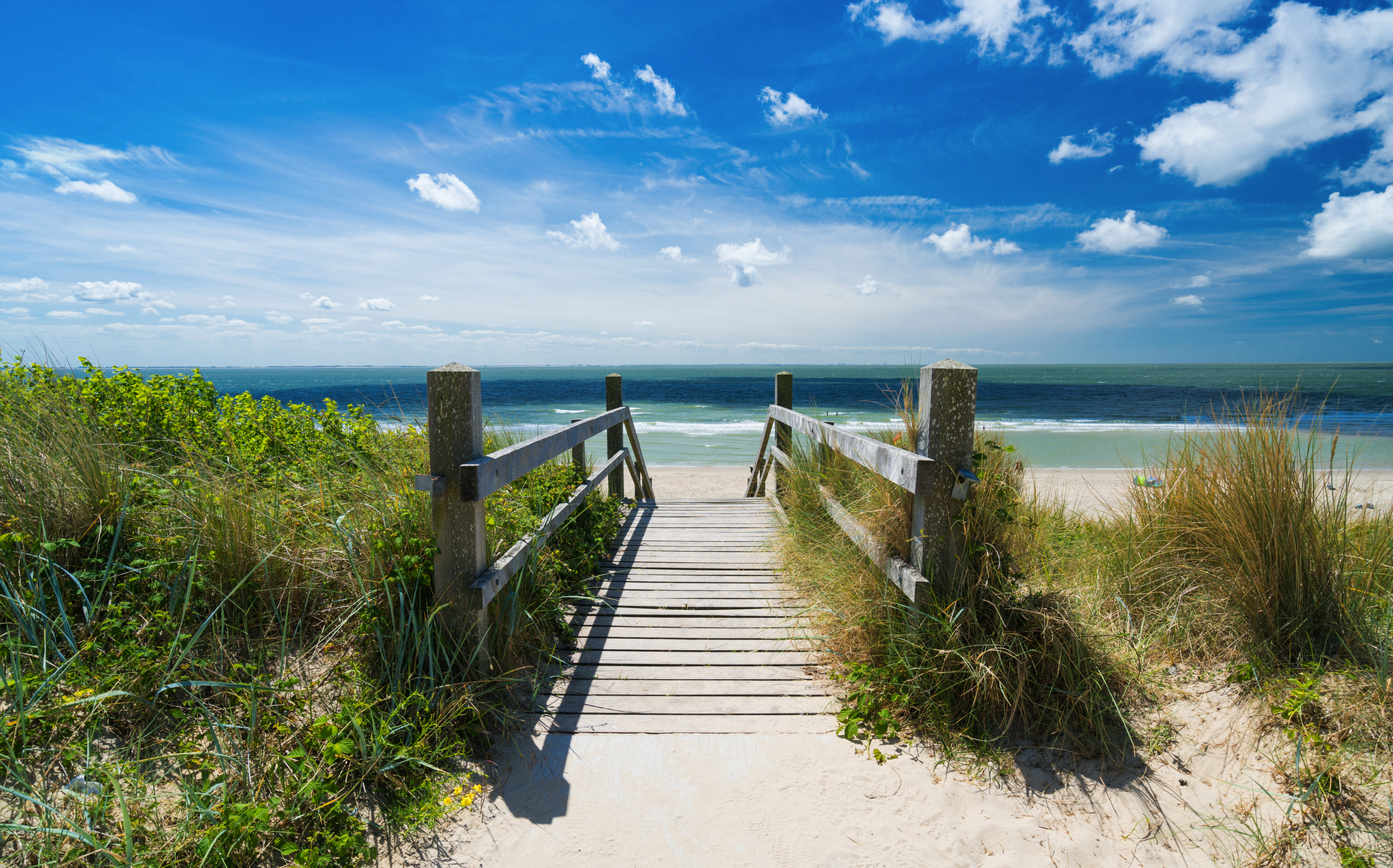 Holzsteg führt durch Dünen zu einem sonnigen Strand mit blauem Himmel und Wolken.