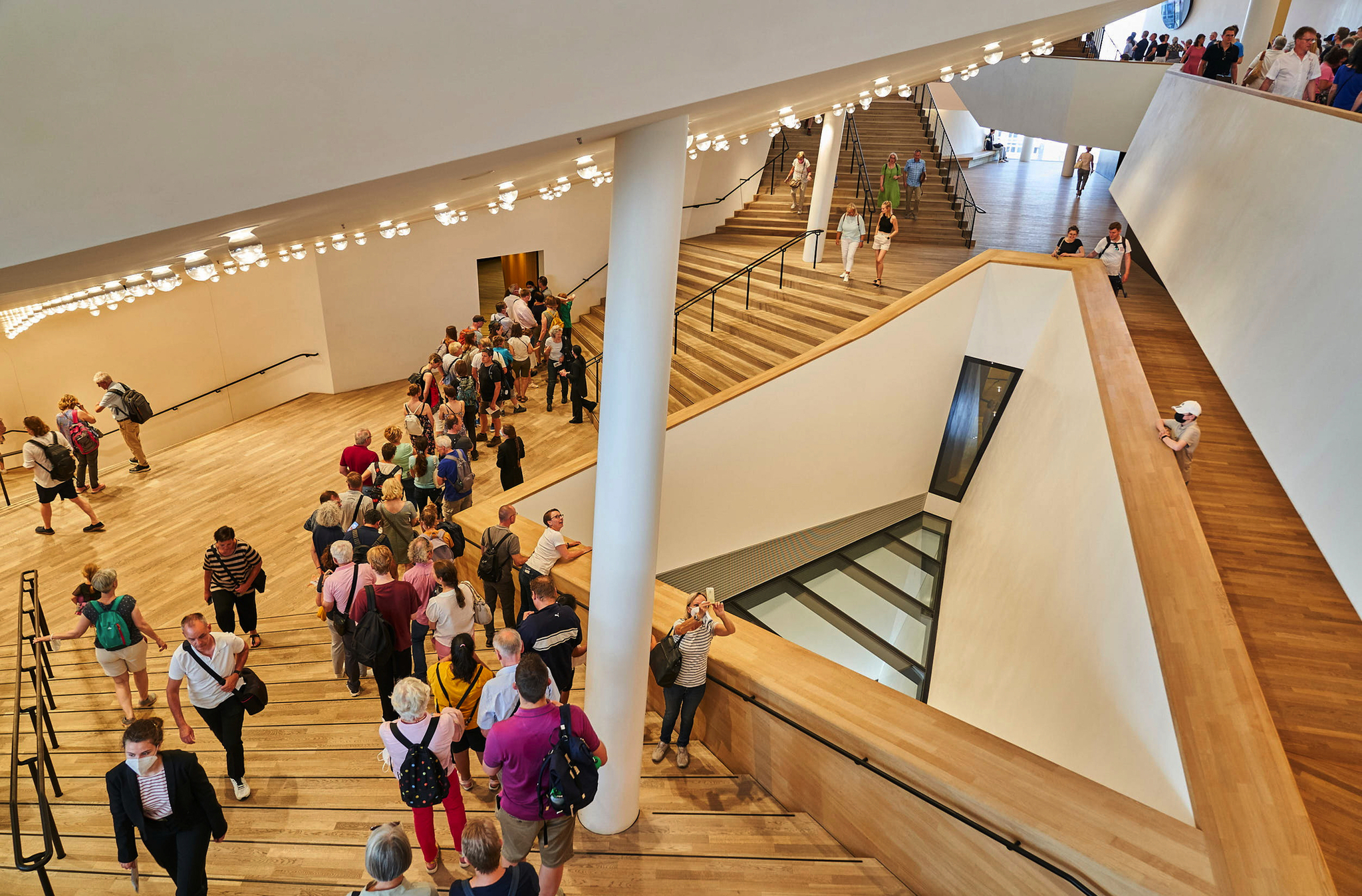 Menschenmenge in einem modernen Gebäude mit Treppen und großen Leuchten.