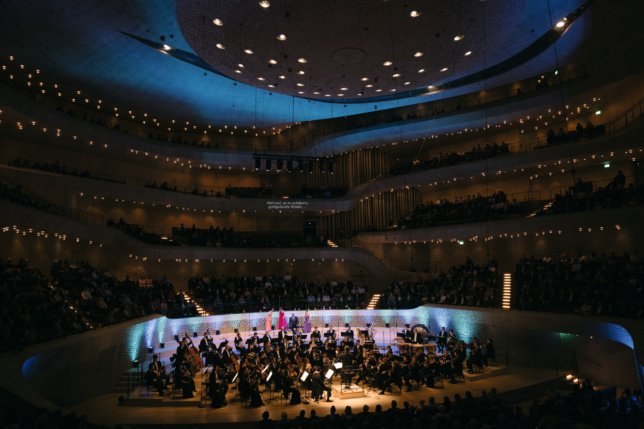 Orchester spielt in Konzertsaal mit beleuchtetem Publikum.