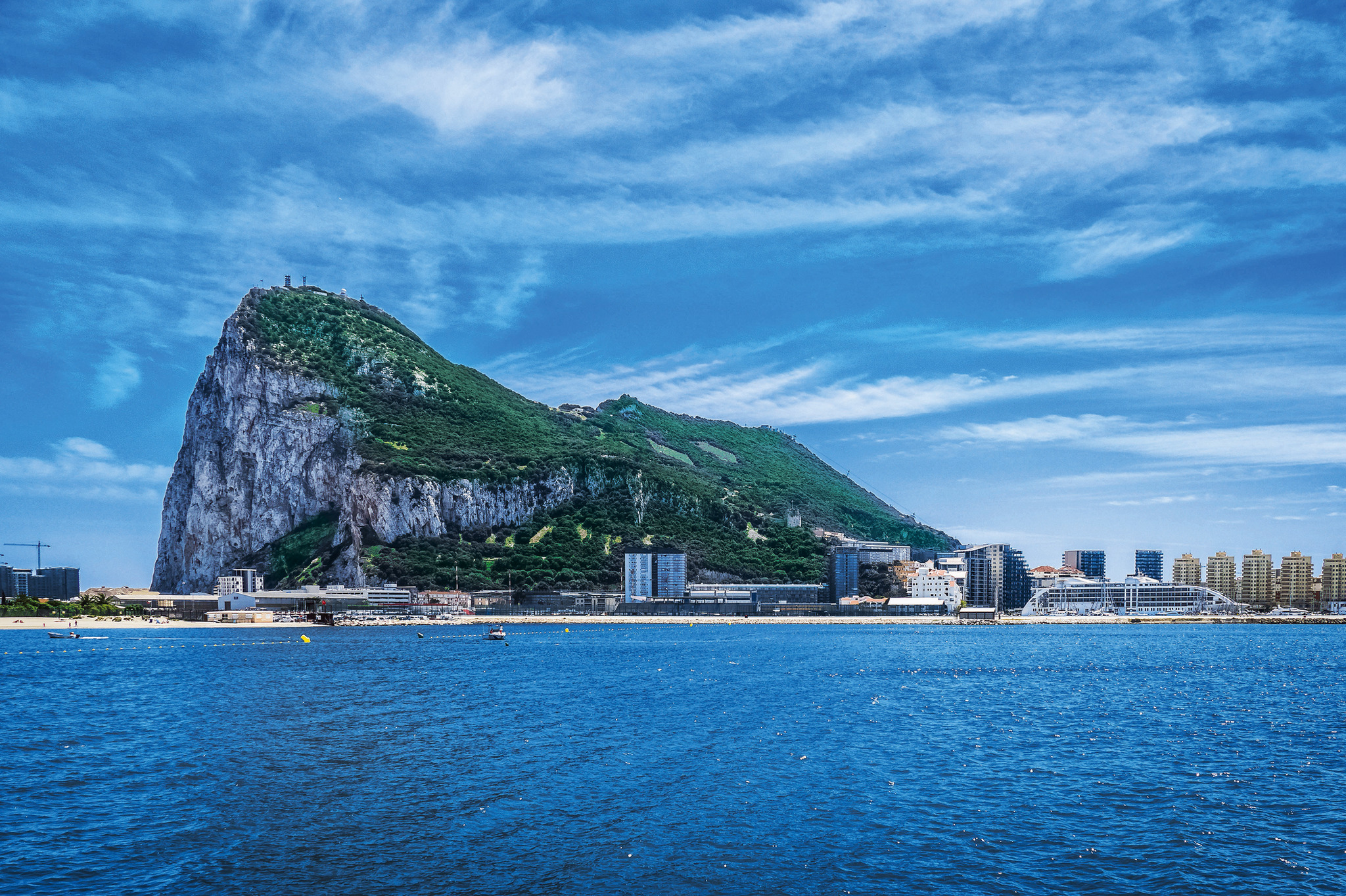 Küste mit Wasser, Gebäuden und einem großen Felsen im Hintergrund unter blauem Himmel
