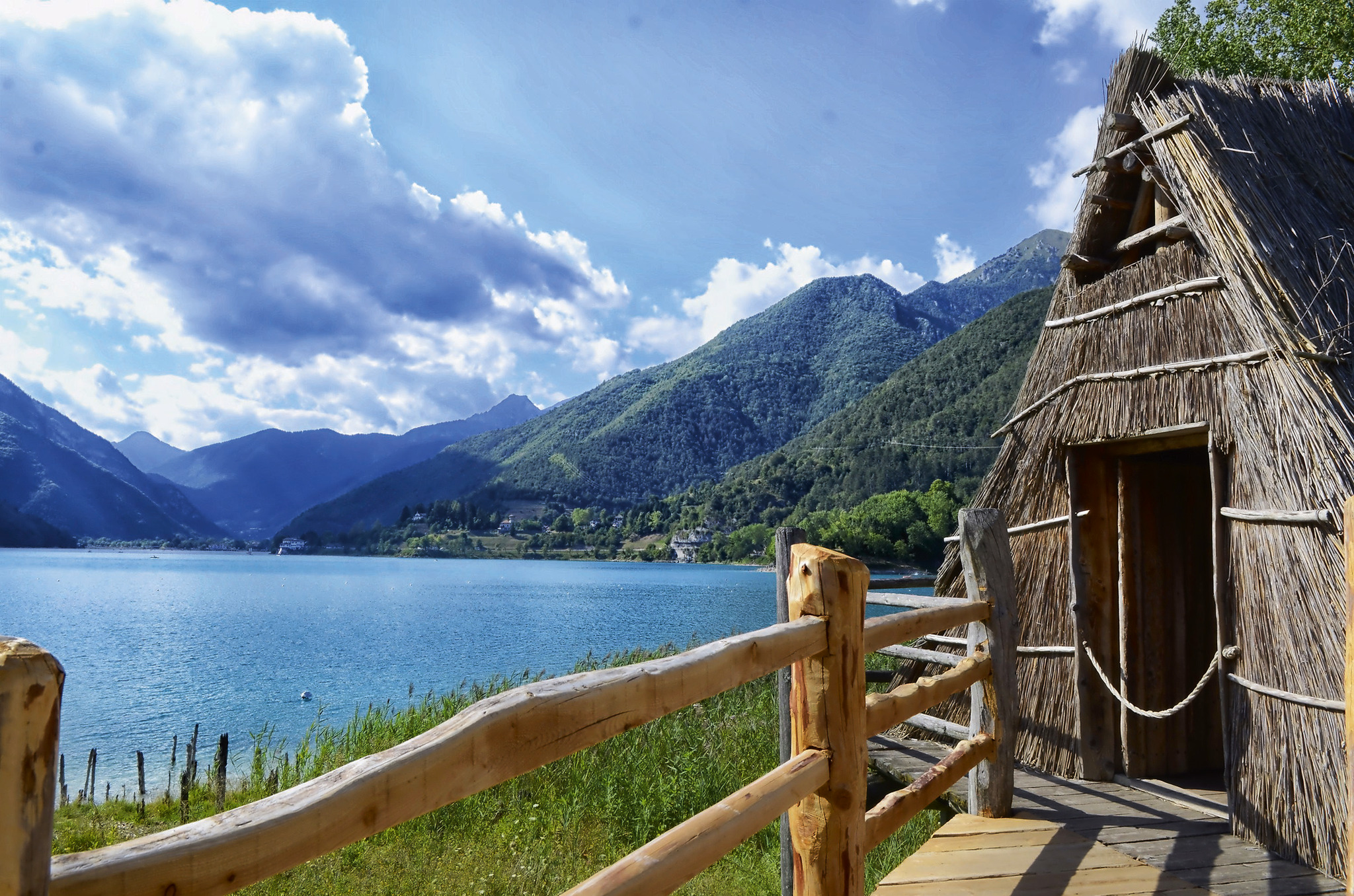 Strohhütte am See mit Bergkulisse und bewölktem Himmel im Hintergrund.
