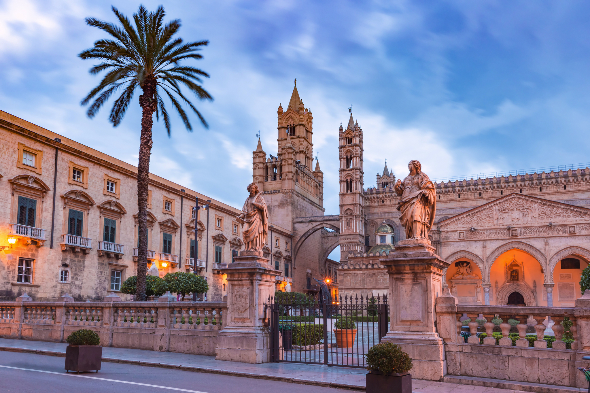 Historische Kathedrale mit Palmen und Statuen im Vordergrund bei Sonnenuntergang.