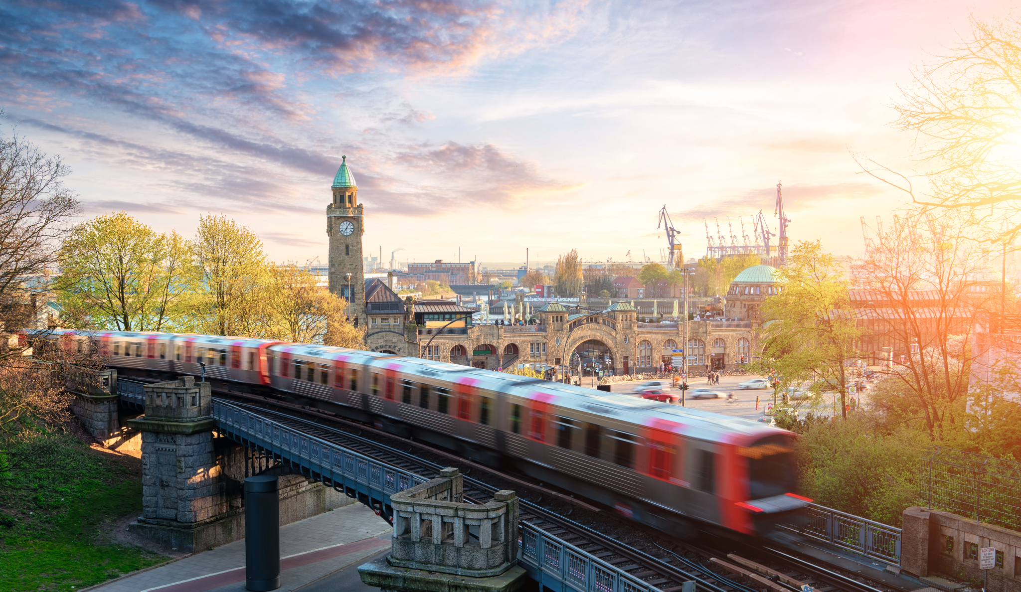 Zug fährt auf Brücke vor Hamburger Hafen und historischem Gebäude bei Sonnenuntergang.