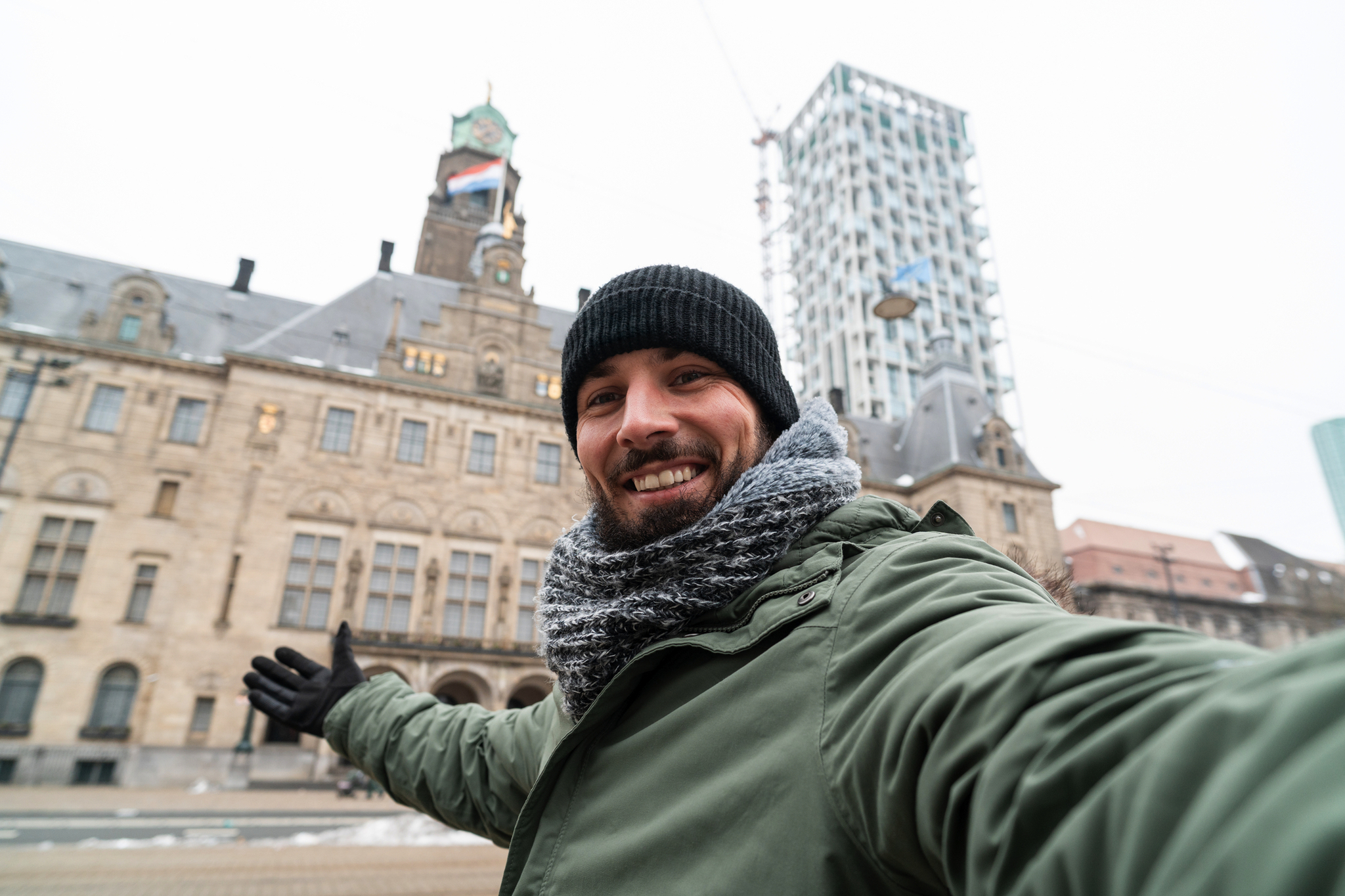 Mann mit Mütze und Schal macht Selfie vor historischem Gebäude und modernem Turm.