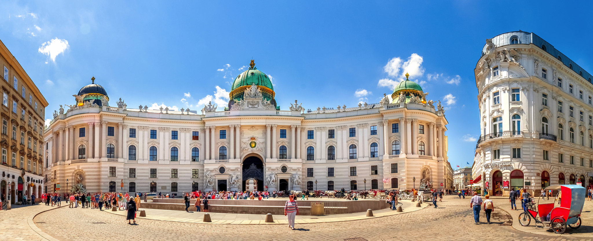 Panorama der Wiener Hofburg mit belebtem Platz und klarer Himmel im Hintergrund.