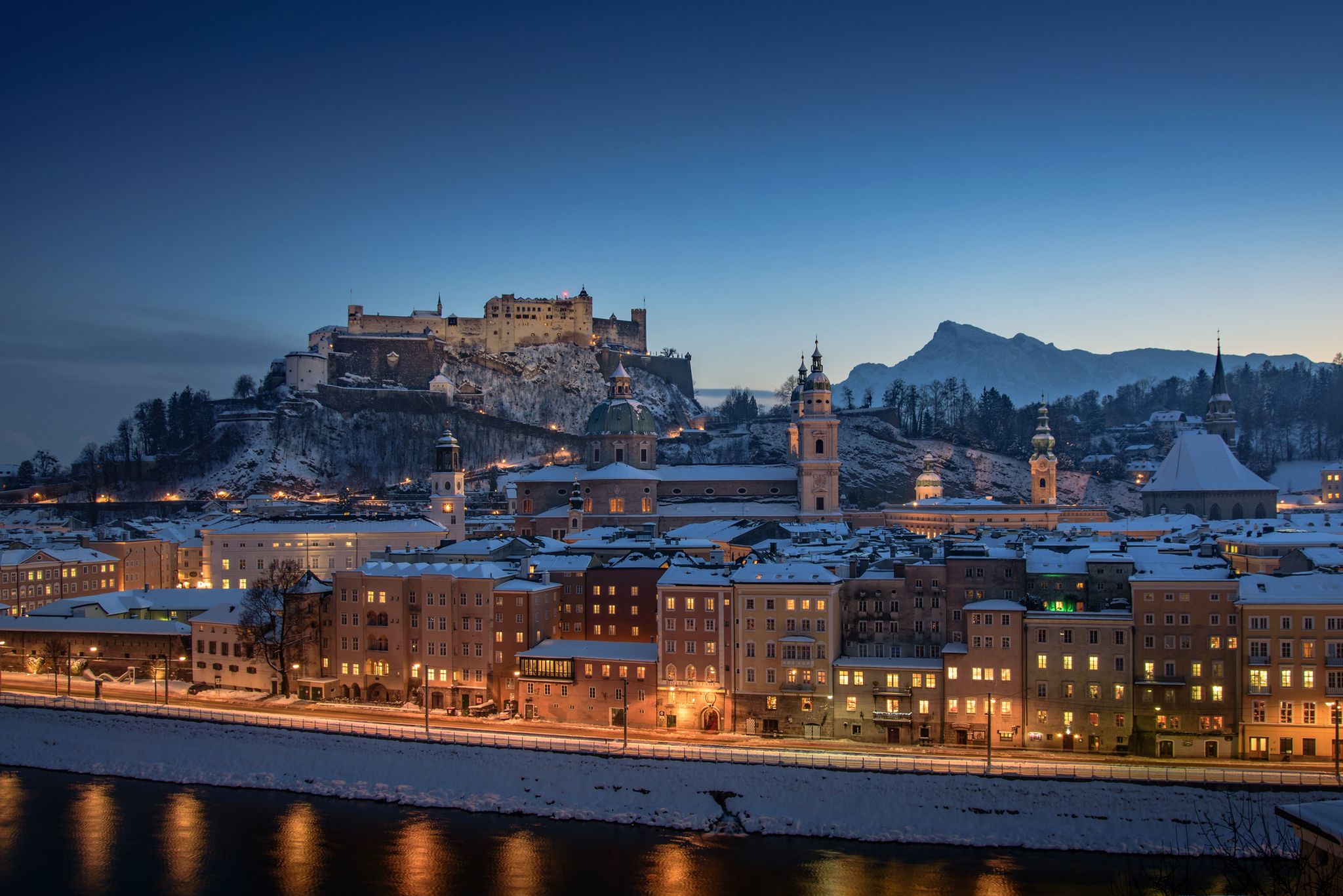 Verschneite Stadt und Burg bei Abenddämmerung, beleuchtet entlang eines Flusses.