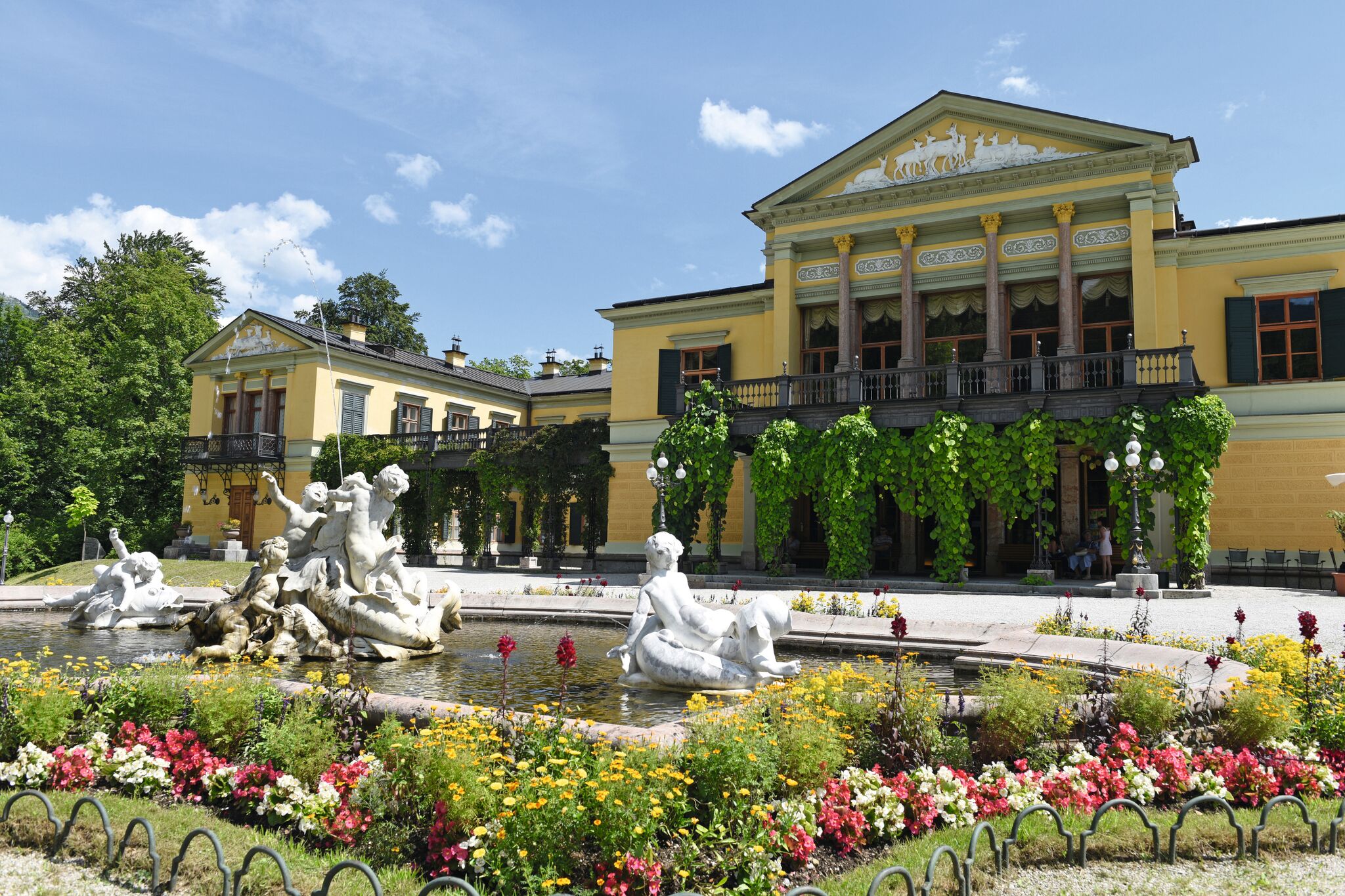 Großes gelbes Gebäude mit Säulen und Brunnen, umgeben von blühendem Garten.