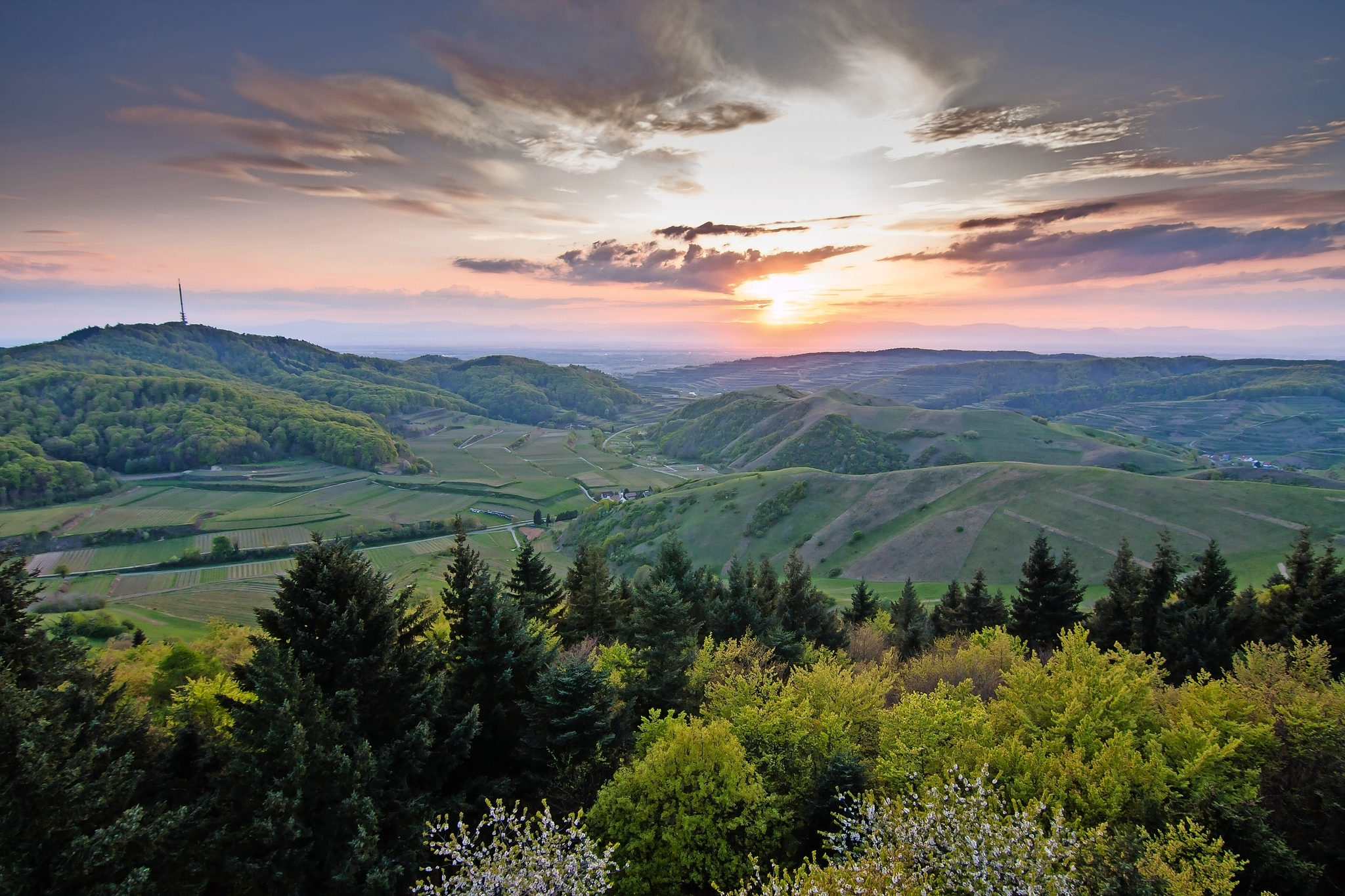 Aussicht über eine hügelige Landschaft mit Wäldern und Sonnenuntergang am Horizont.