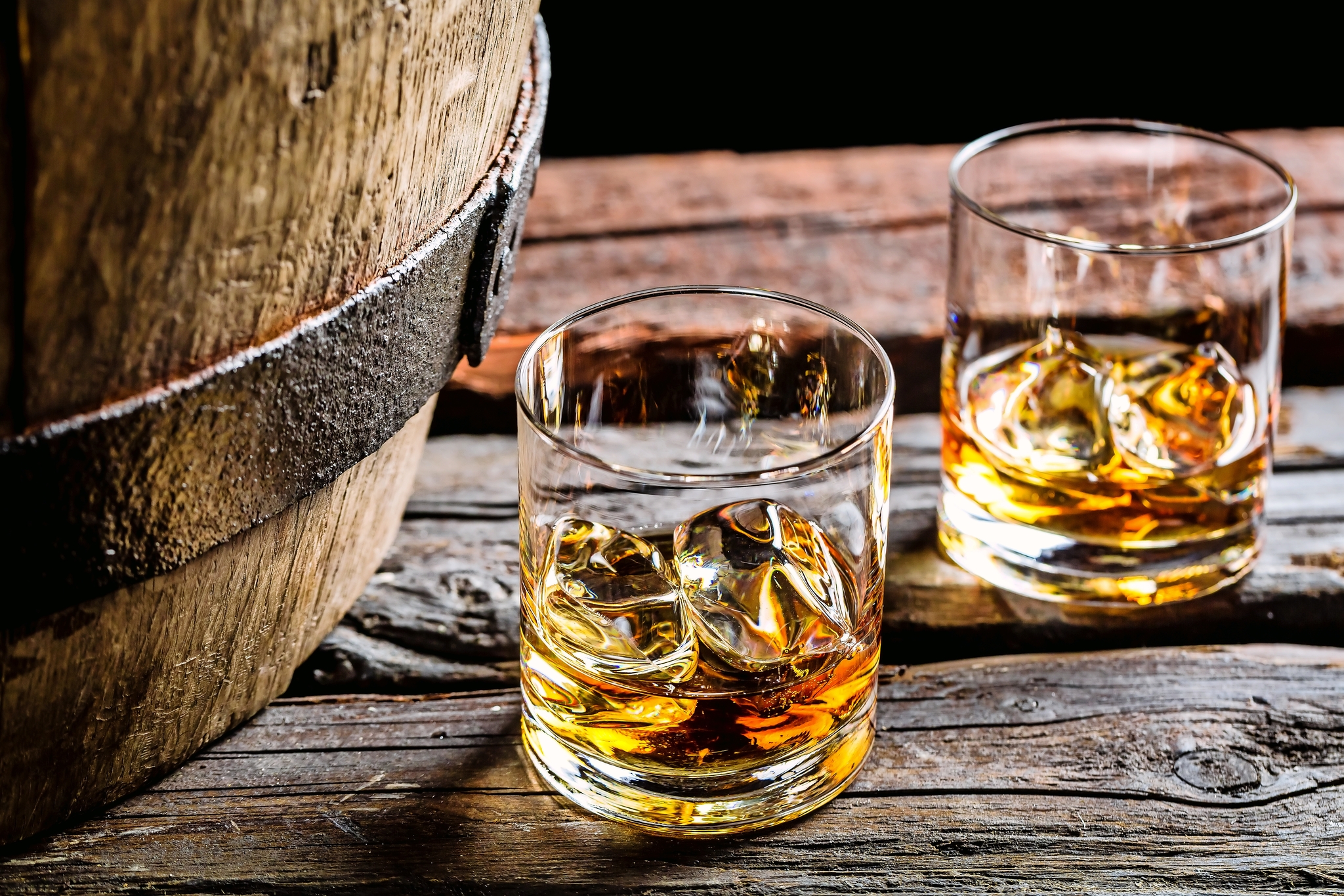 Zwei Whiskygläser mit Eis auf einem Holztisch neben einem Fass.