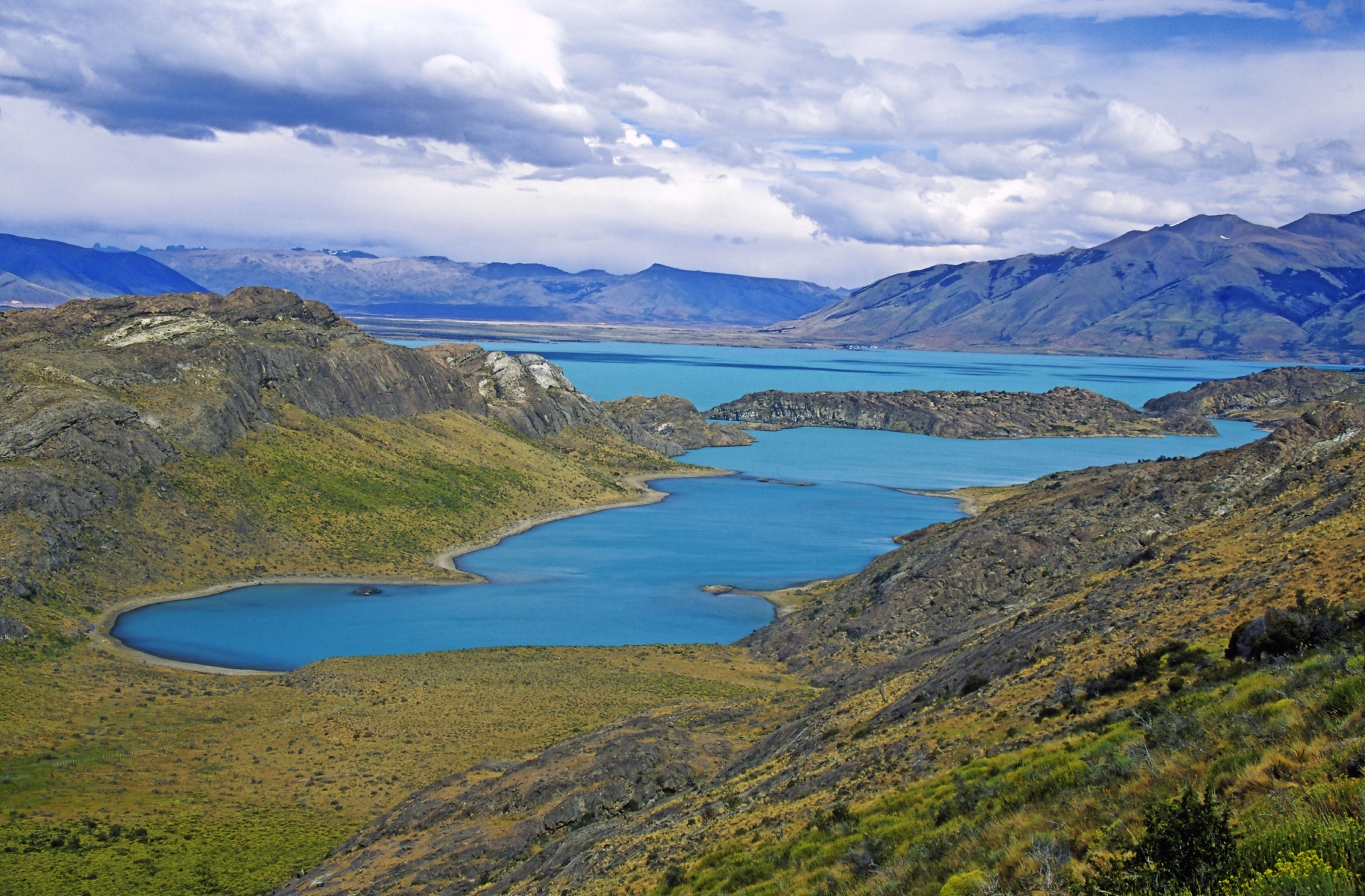 Blick auf eine Landschaft mit blauem See und umliegenden Bergen unter bewölktem Himmel.