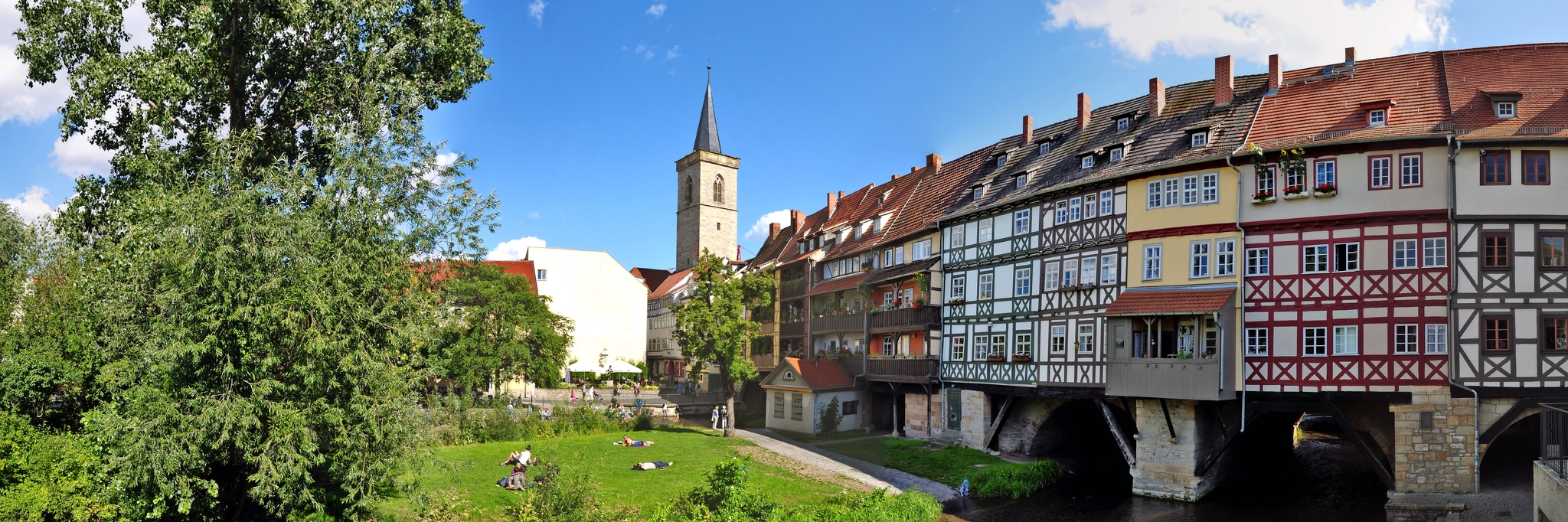 Brücke mit Fachwerkhäusern und Kirchturm, grüner Park im Vordergrund.