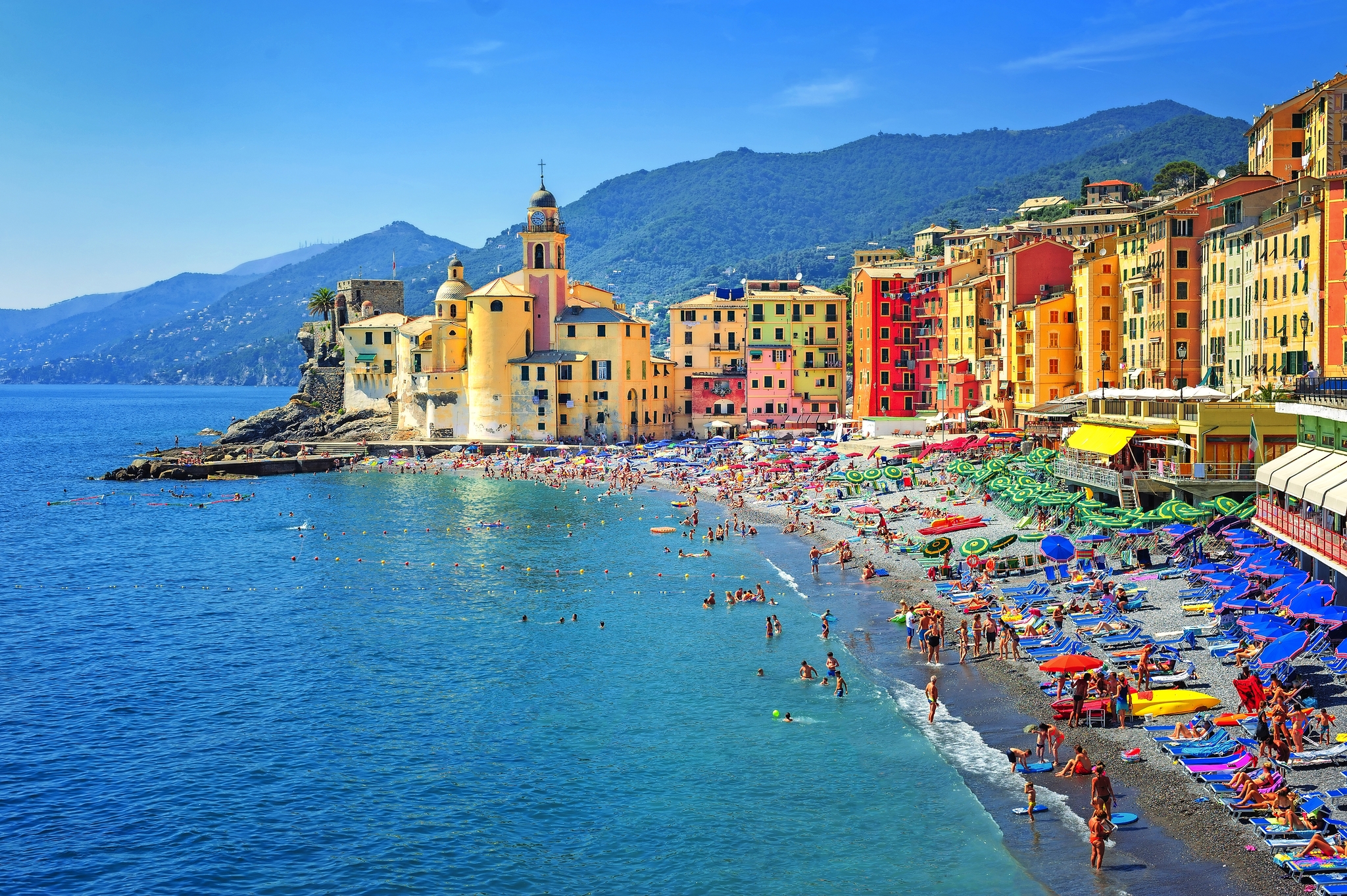 Bunte Küstenstadt mit Strand, zahlreichen Badenden und bunten Sonnenschirmen.