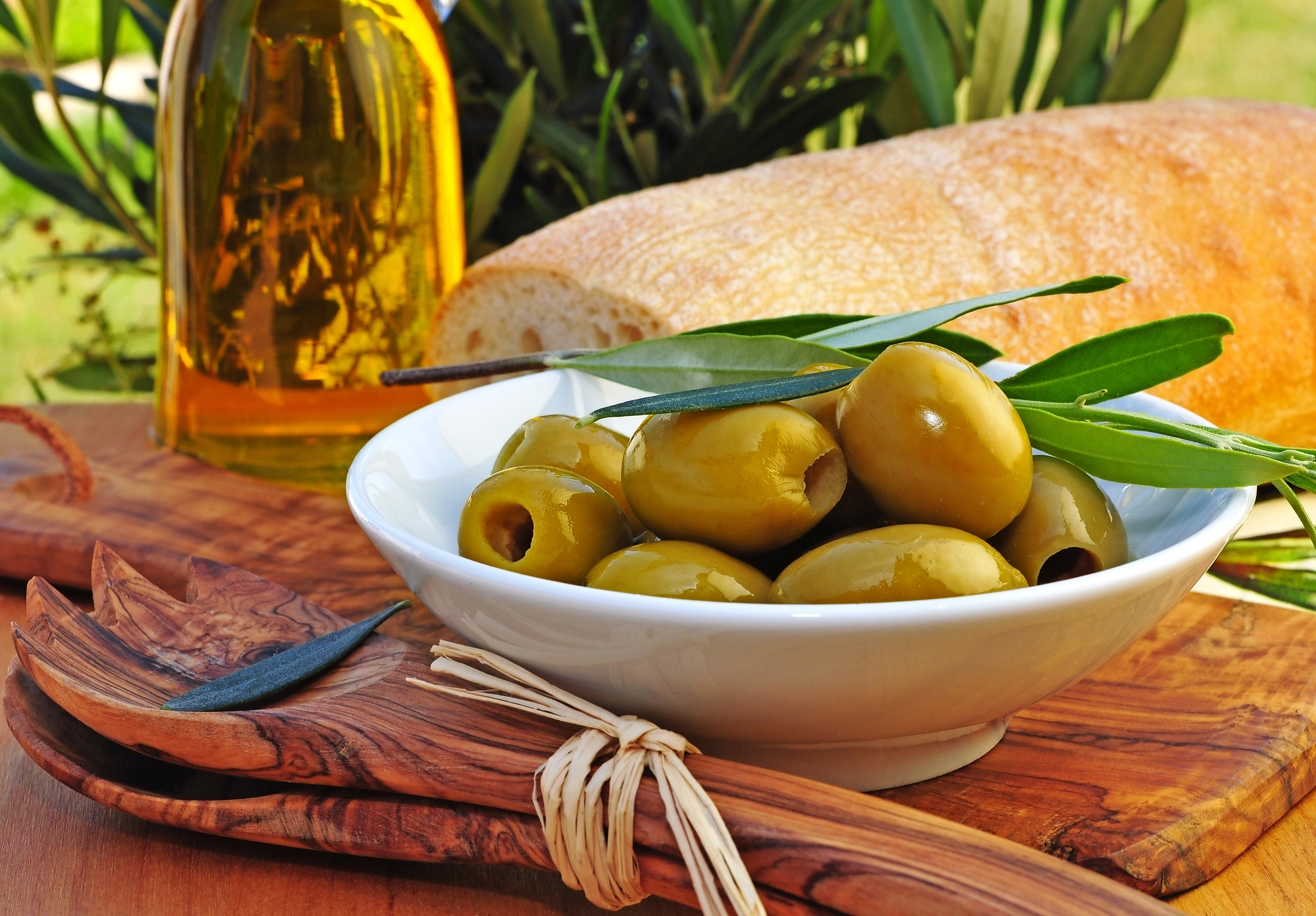 Schale mit grünen Oliven, Olivenölflasche, Holzlöffel, frisches Brot im Hintergrund.