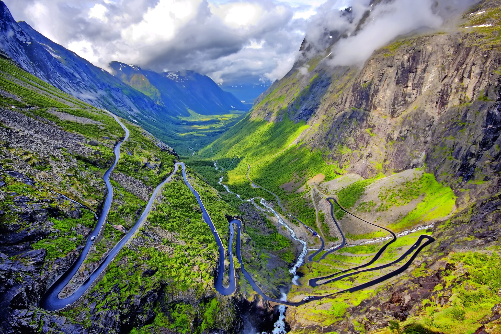 Serpentinenstraße in einem grünen, bergigen Tal mit dramatischem Himmel.