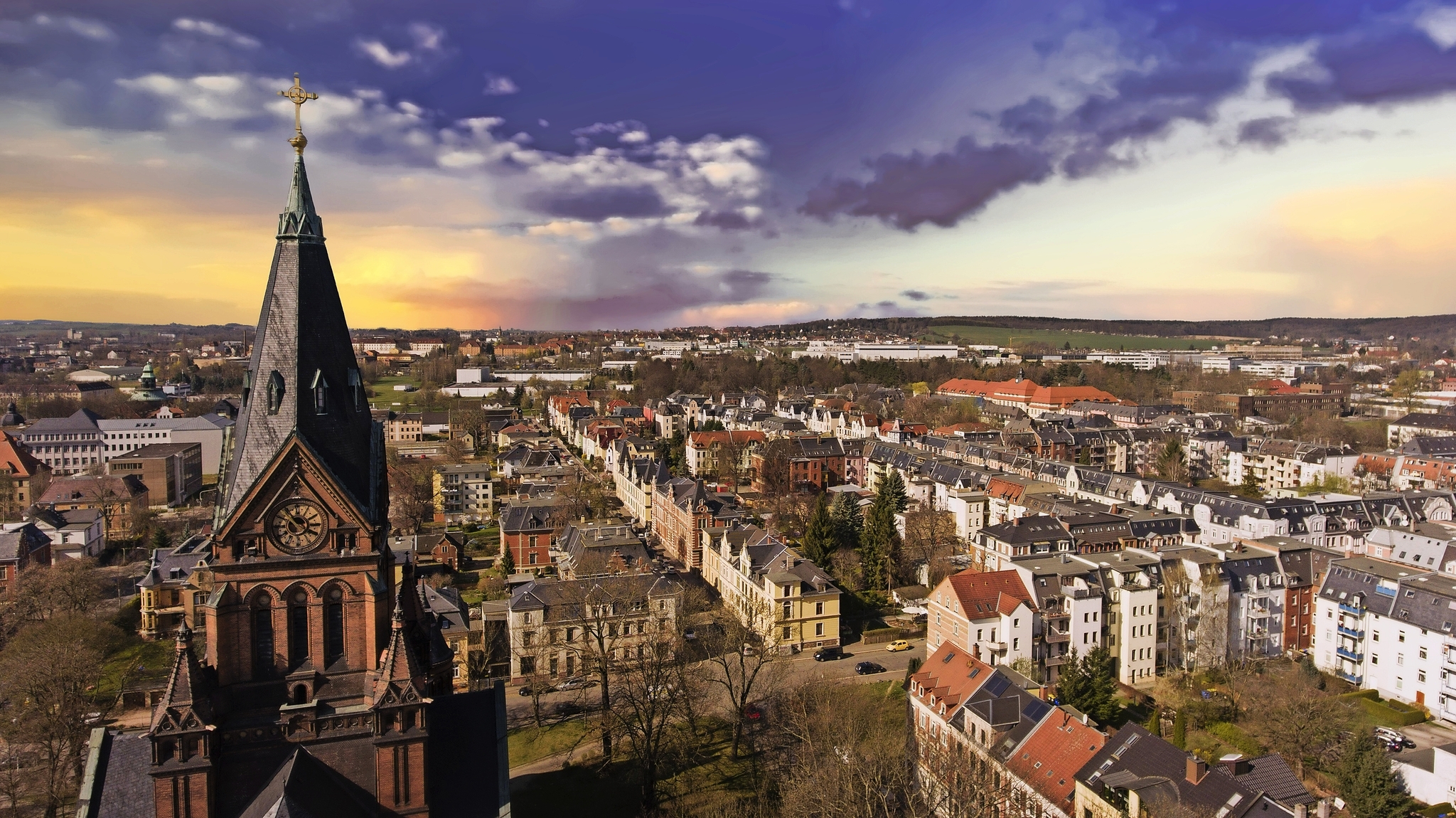 Stadtansicht mit Kirchturm im Vordergrund und buntem Sonnenuntergang.