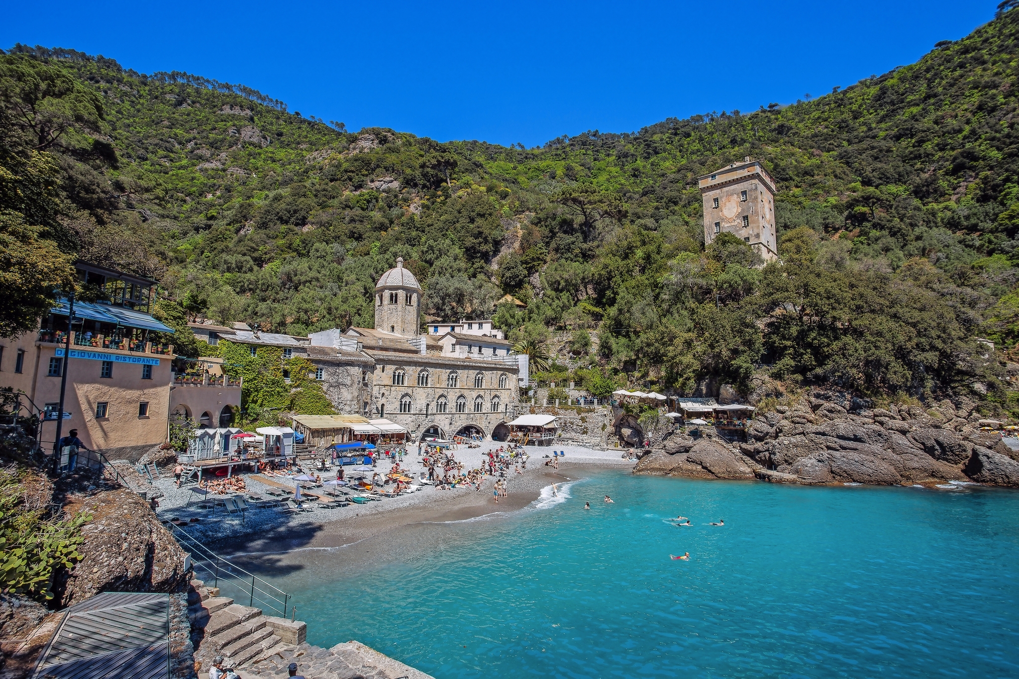 Strand vor einer Klosteranlage mit bewaldeten Hügeln im Hintergrund.