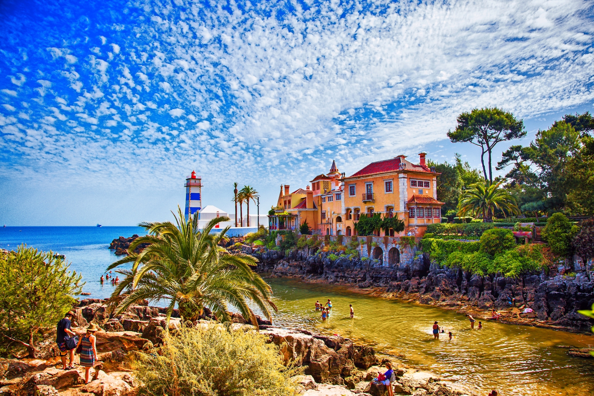 Küstenlandschaft mit Leuchtturm, Villa, Palmen und Menschen am Ufer unter blauem Himmel.