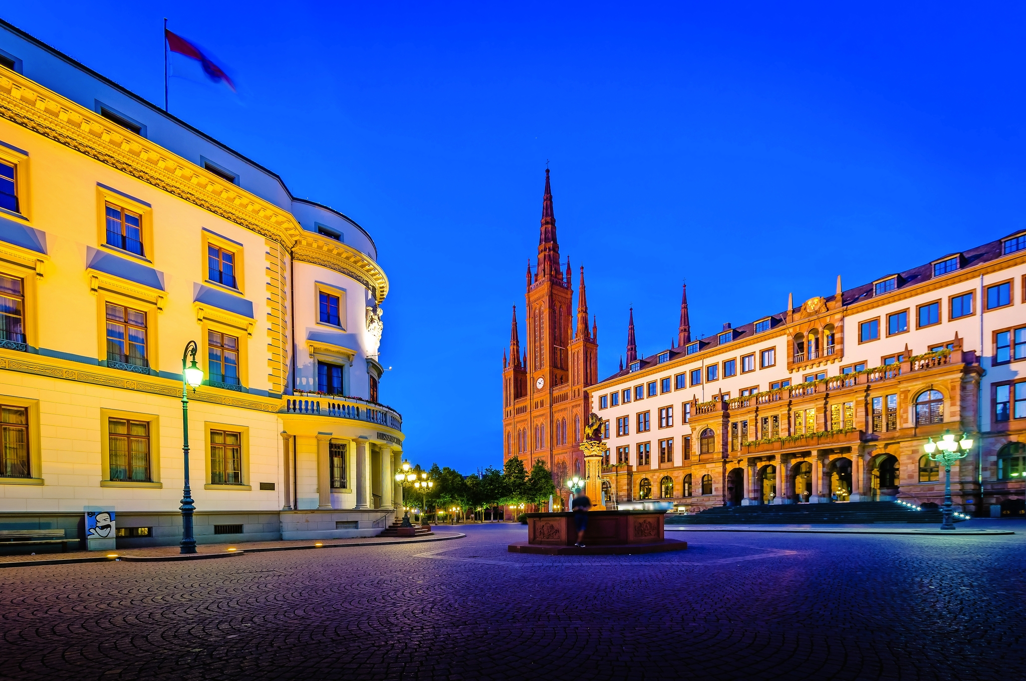 Stadtplatz bei Nacht mit historischer Architektur, links ein beleuchtetes Gebäude mit Flagge, im Hintergrund eine gotische Kirche mit hohen Türmen, umgeben von weiteren historischen Gebäuden.