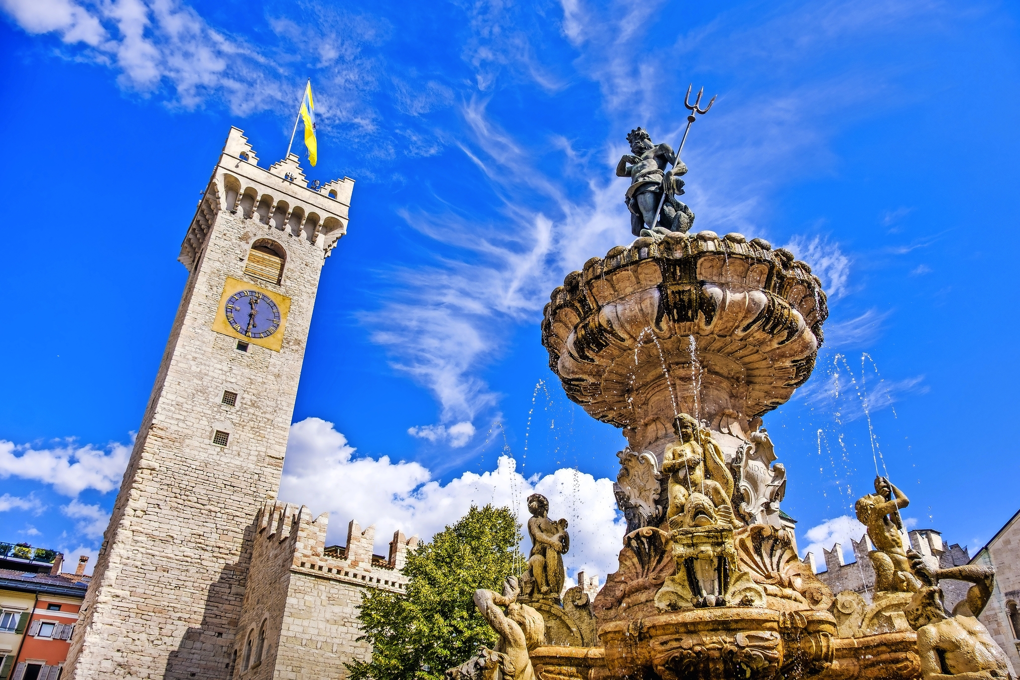 Springbrunnen mit Statuen und Uhrturm im Hintergrund bei blauem Himmel.