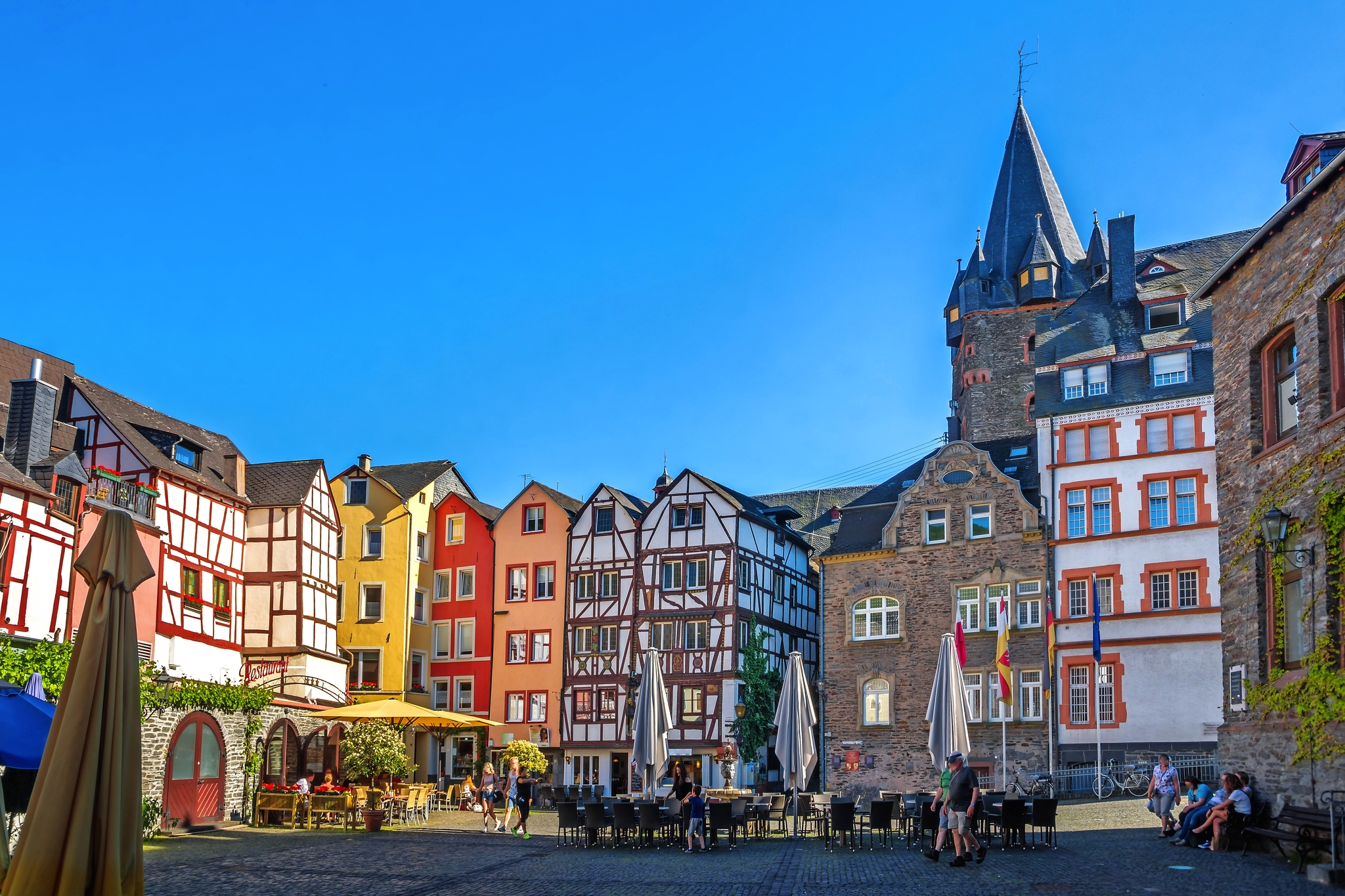 Buntem Fachwerkhäusern umgebener Platz mit Tischen und Sonnenschirmen bei blauem Himmel.