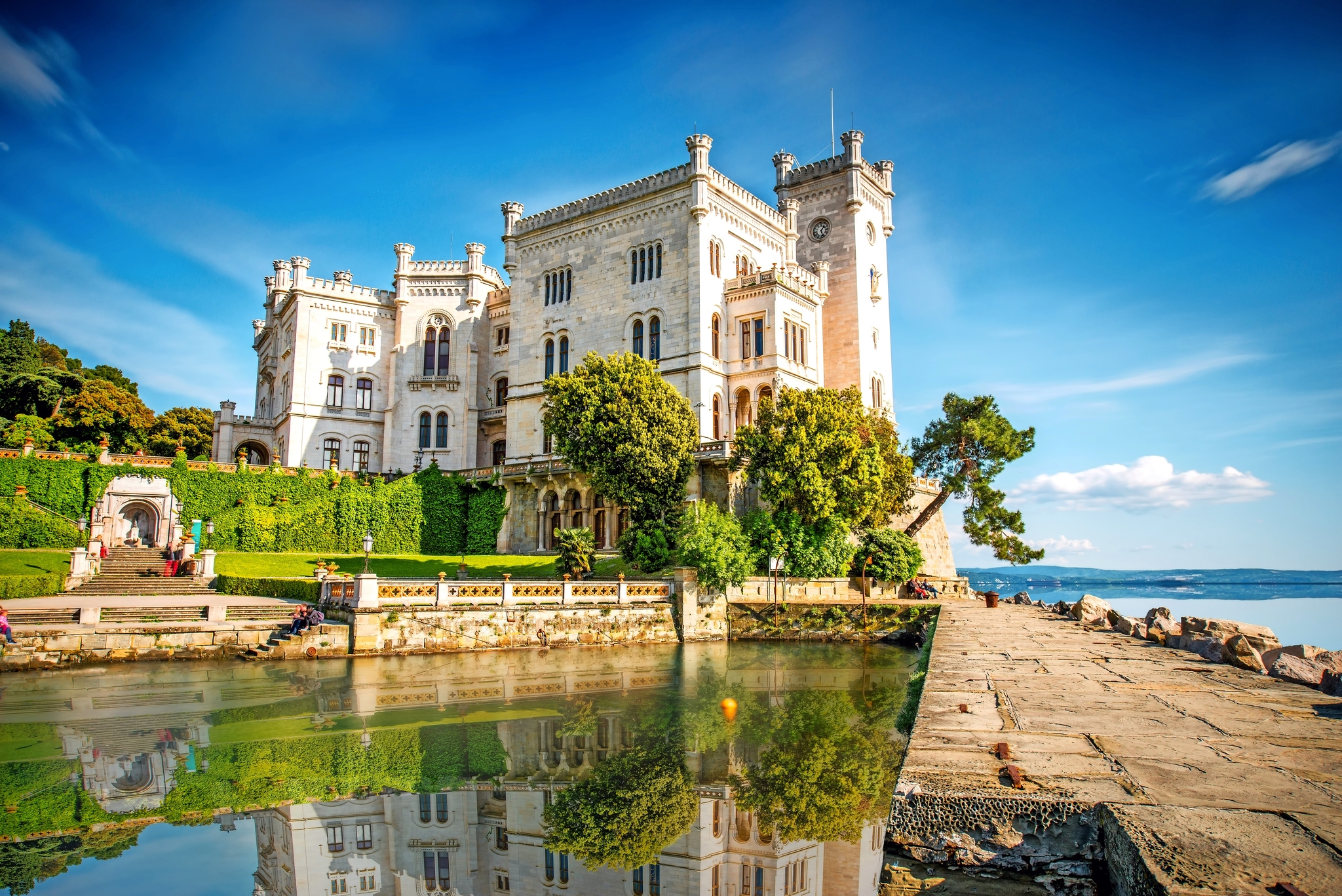 Historisches Schloss am Ufer mit blauem Himmel und reflektierendem Wasser.