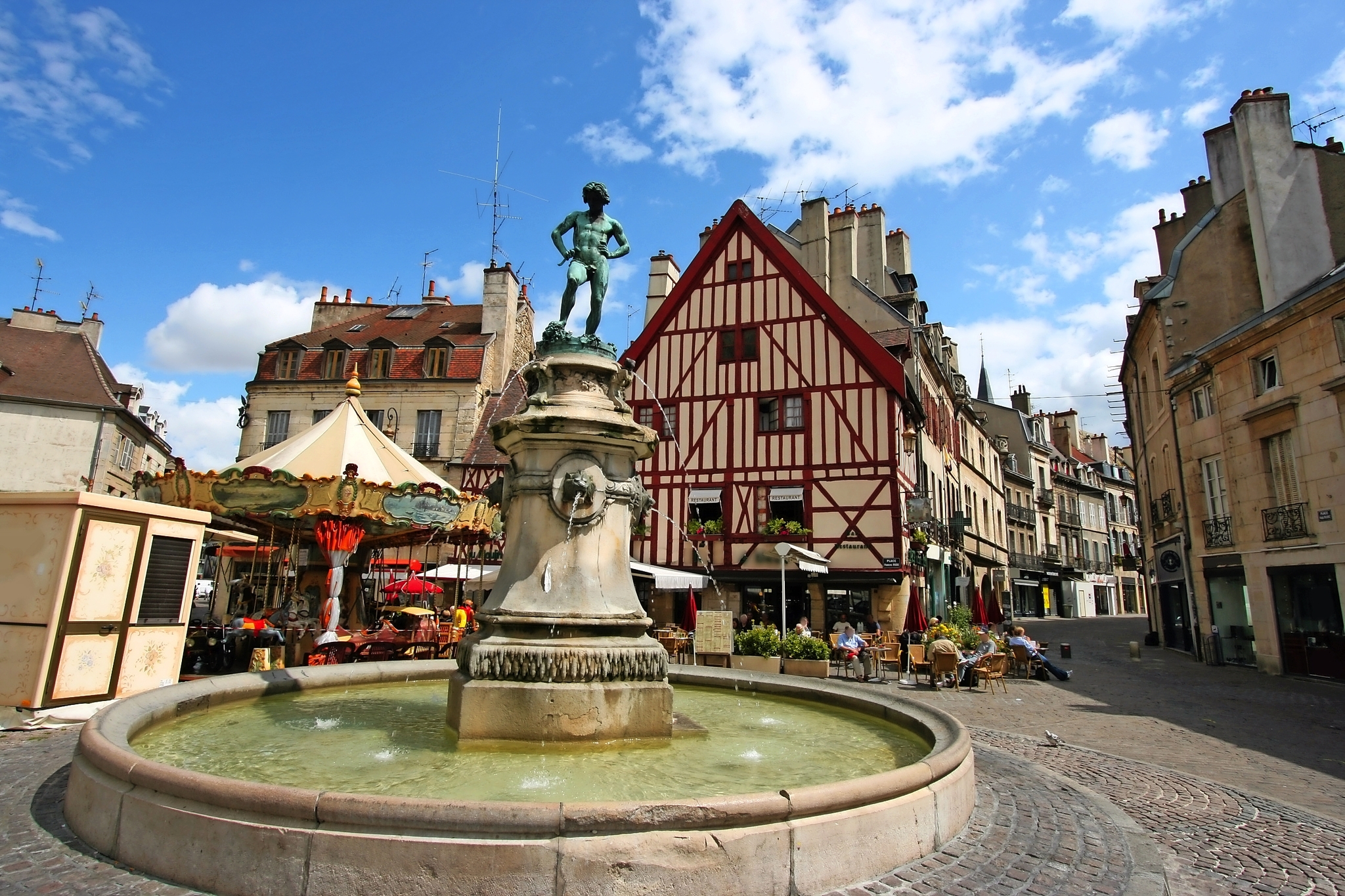 Brunnen auf einem historischen Platz mit Fachwerkhaus, Karussell und blauen Himmel im Hintergrund.