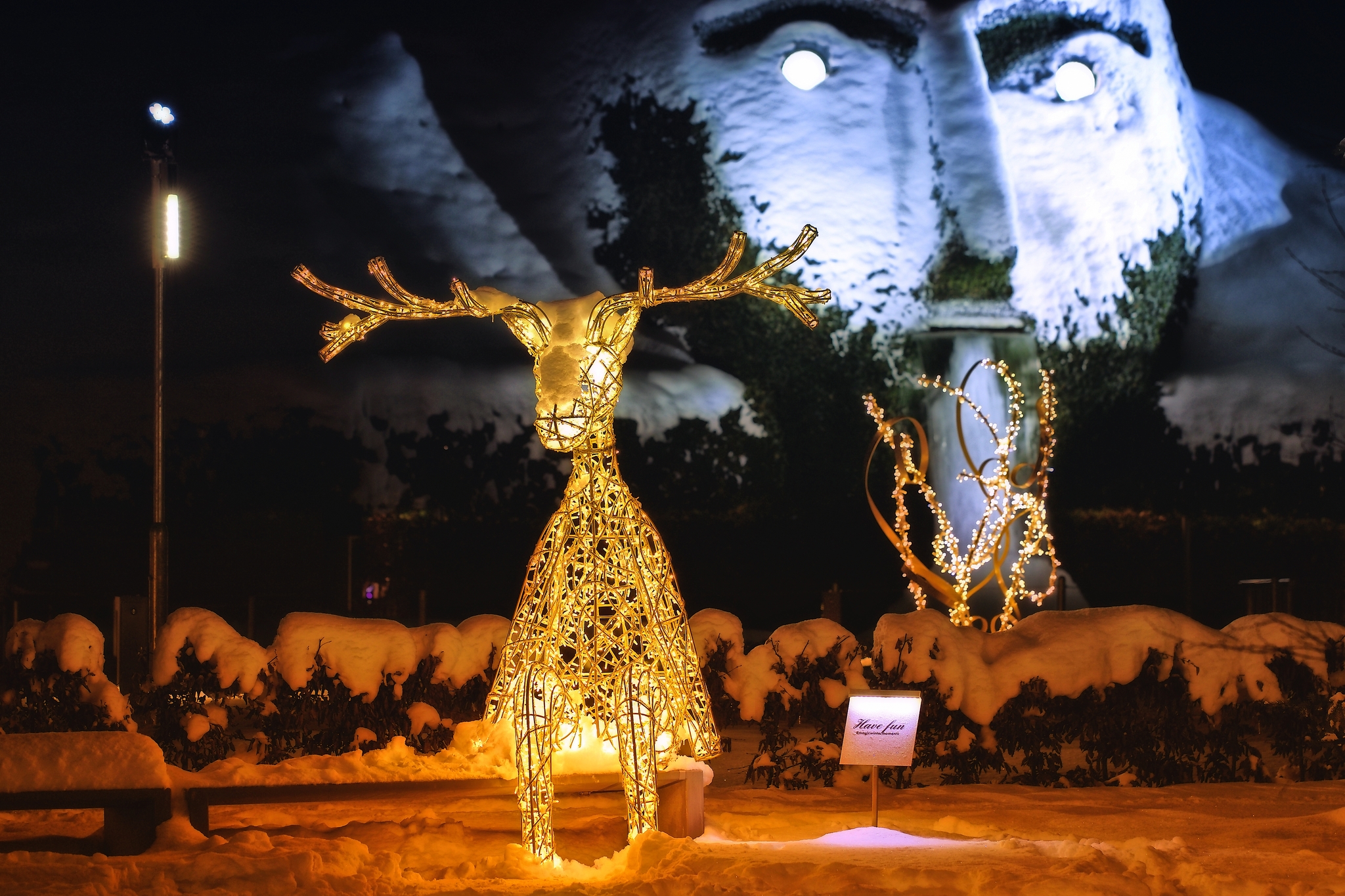 Leuchtende Rentierskulptur und riesiger Gesichtskopf im Schnee bei Nacht.